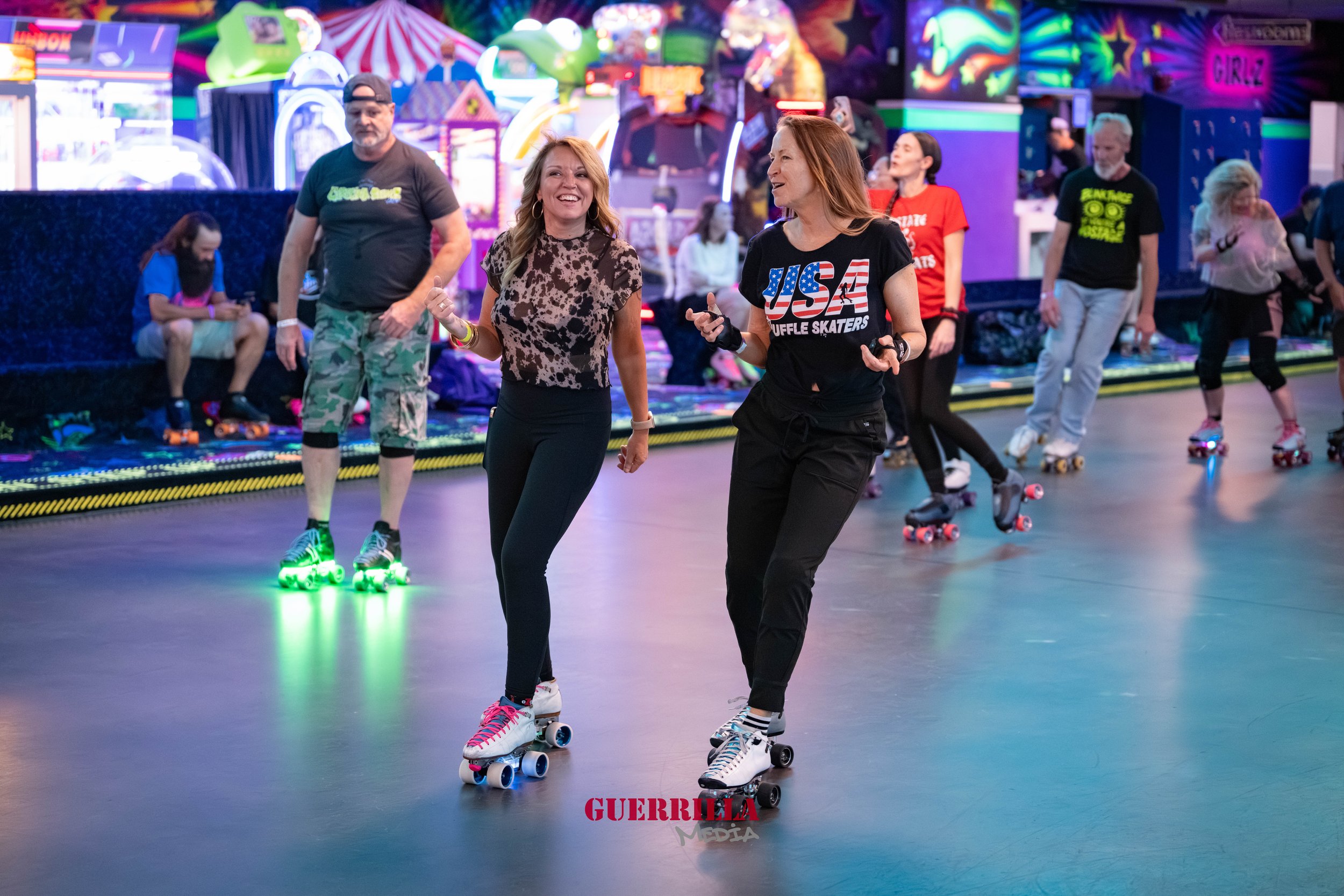 Two women roller skating and smiling at an indoor roller rink with colorful arcade games and other skaters in the background.