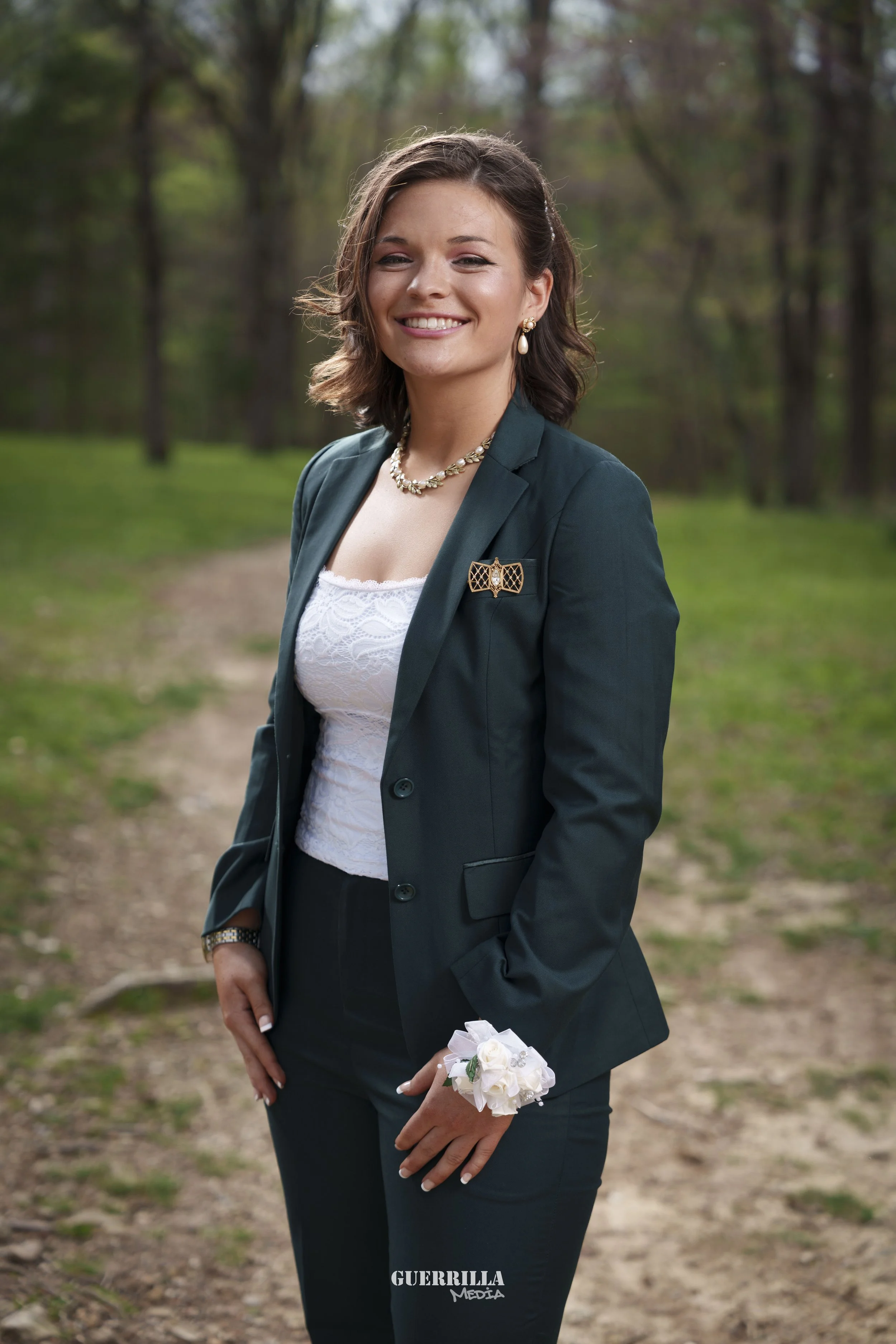 A woman dressed in a suit with a necklace, earrings, and a corsage standing on a wooded path outdoors, smiling at the camera.