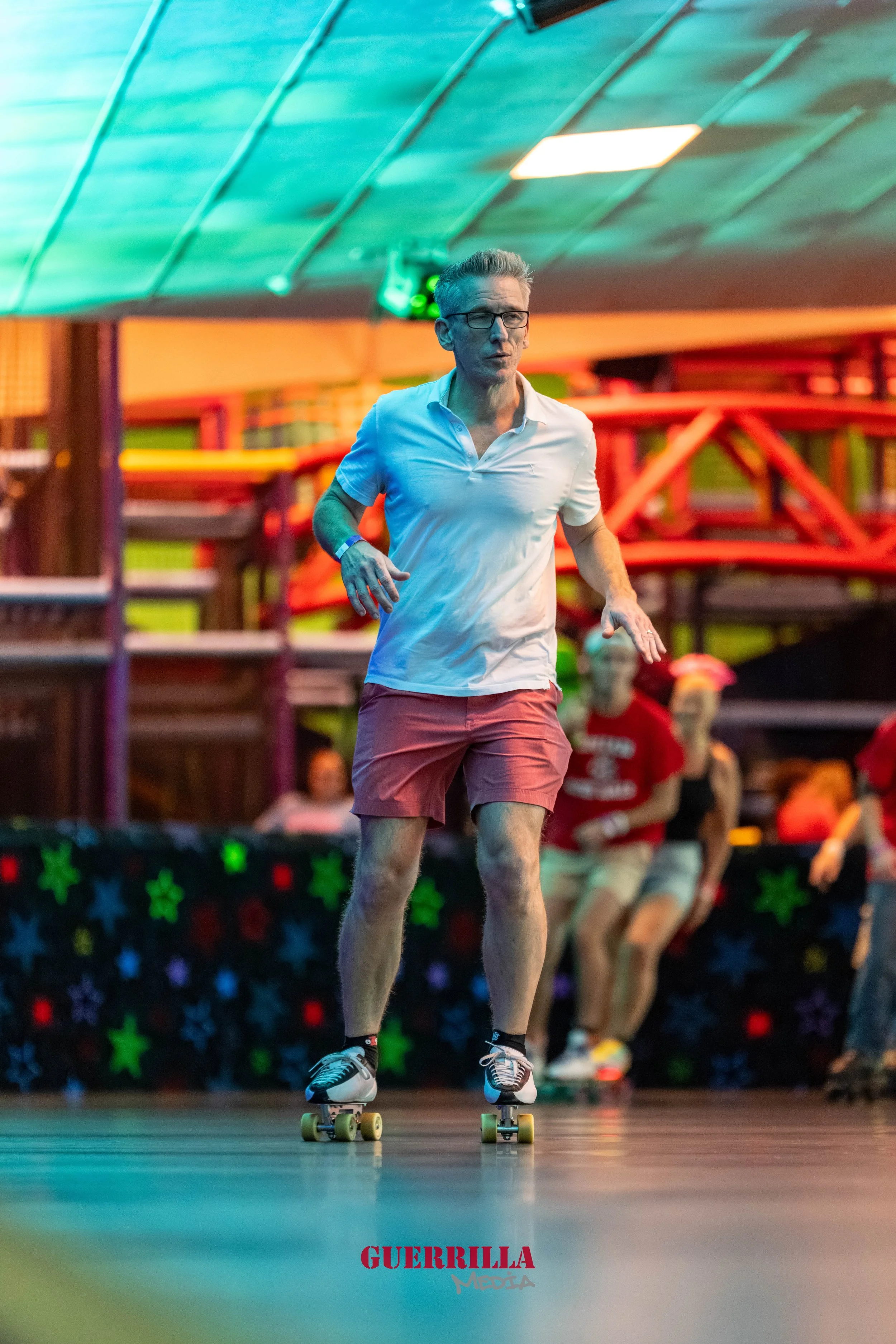 A man roller skating indoors with colorful lighting, wearing a white polo shirt and red shorts, with people and colorful structures in the background.