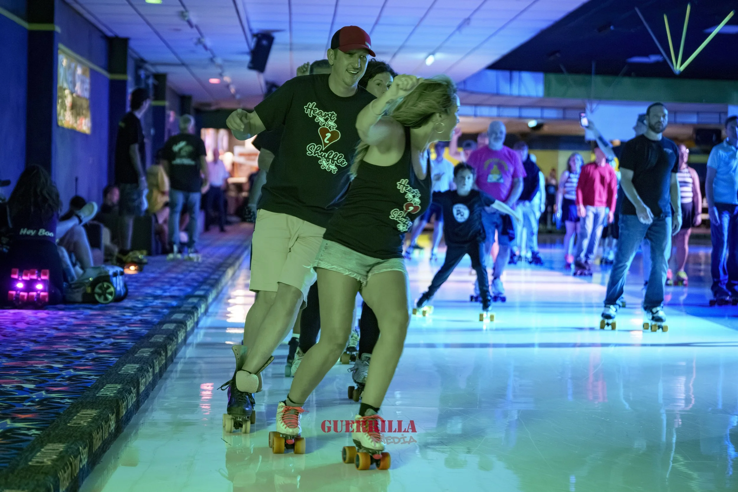 People roller skating indoors under colorful lighting, with some wearing matching black T-shirts with the phrase 'Heart Shuffle' and a heart logo.