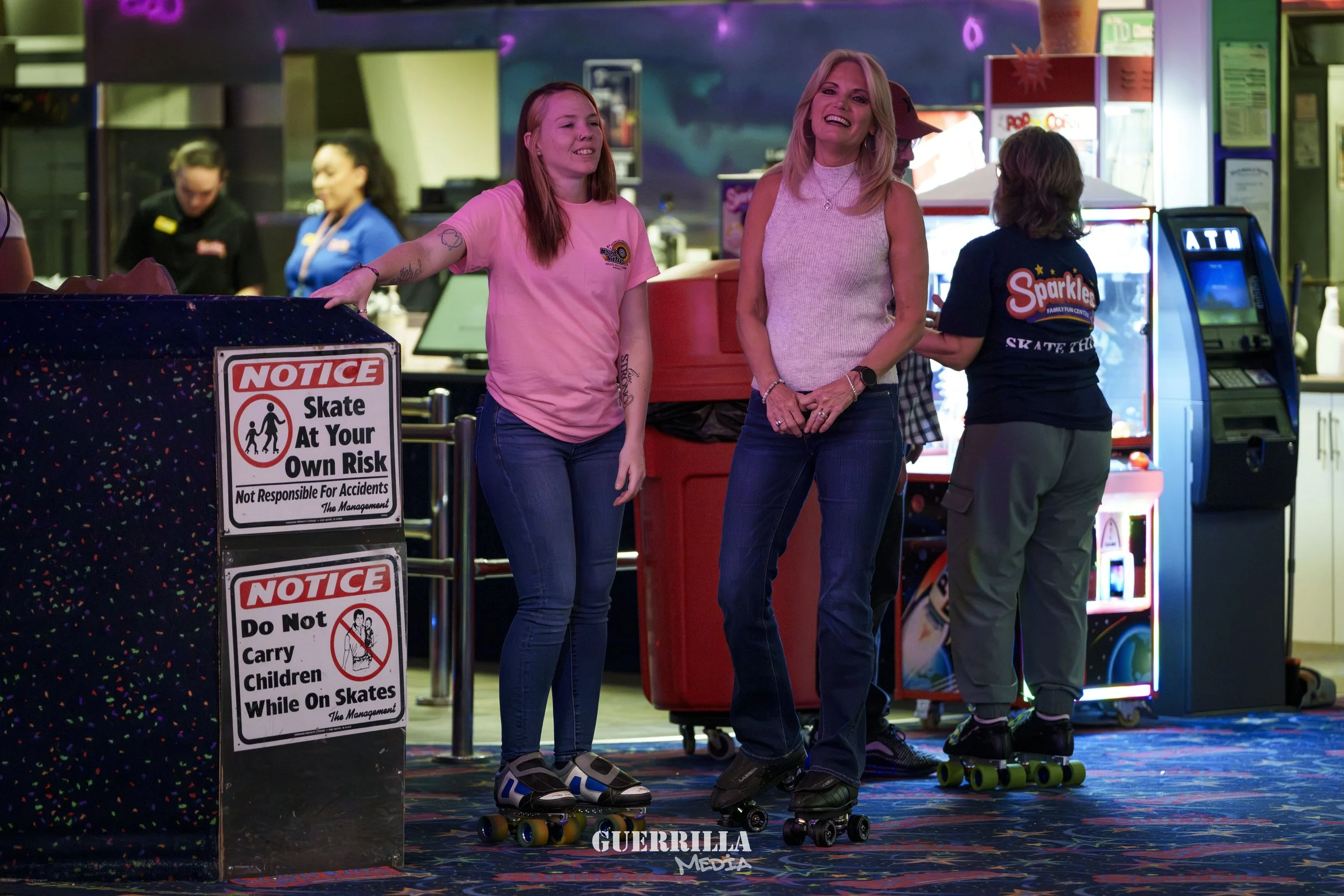 Two women in an indoor skating rink or arcade, one young and one older, both on roller skates and smiling, with signs warning about skating at your own risk and not carrying children while on skates. Other people are visible in the background.