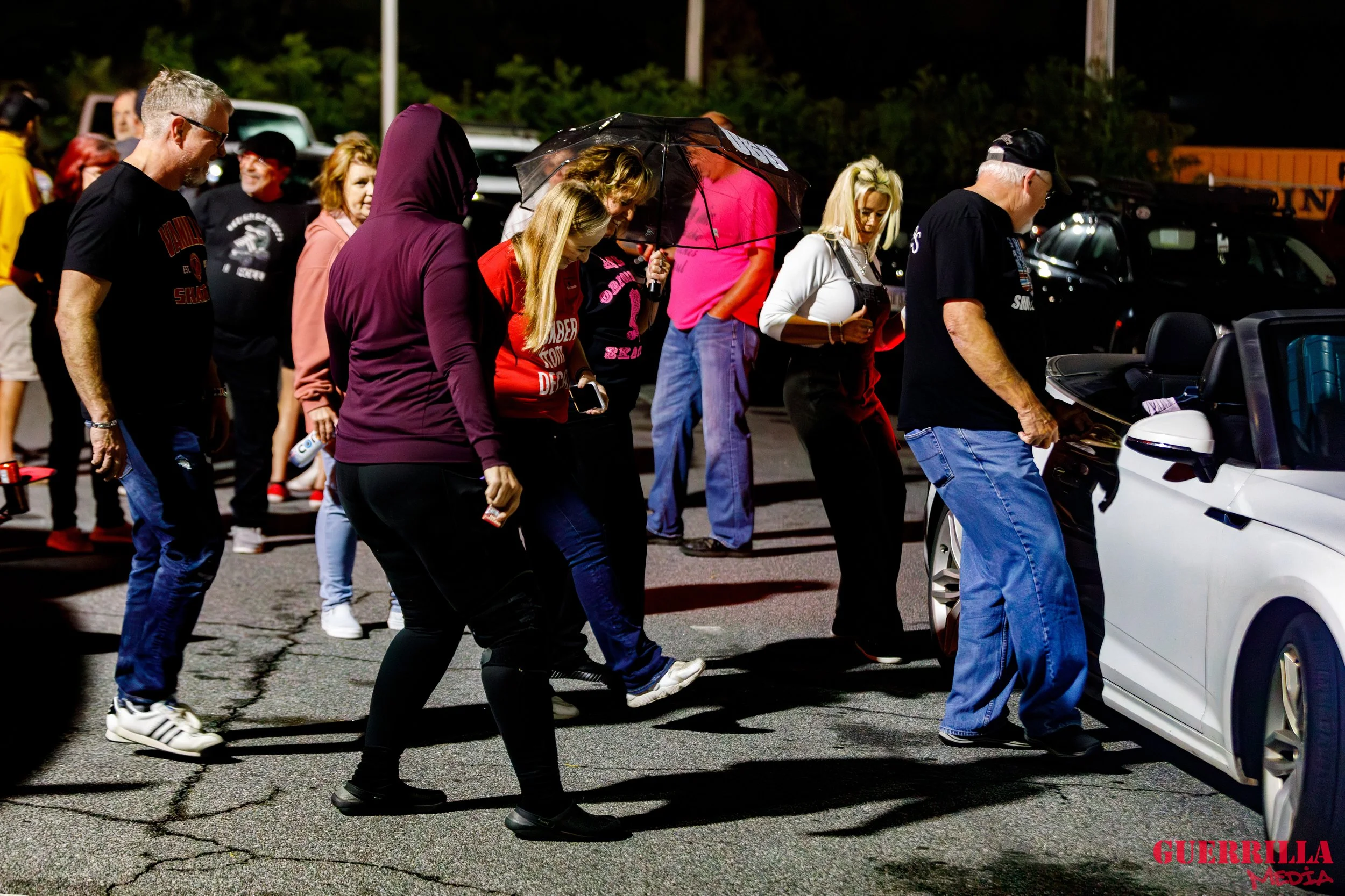 People standing and looking at a white convertible car in a parking lot at night, some people are taking photos, one person is holding an umbrella, others are chatting.