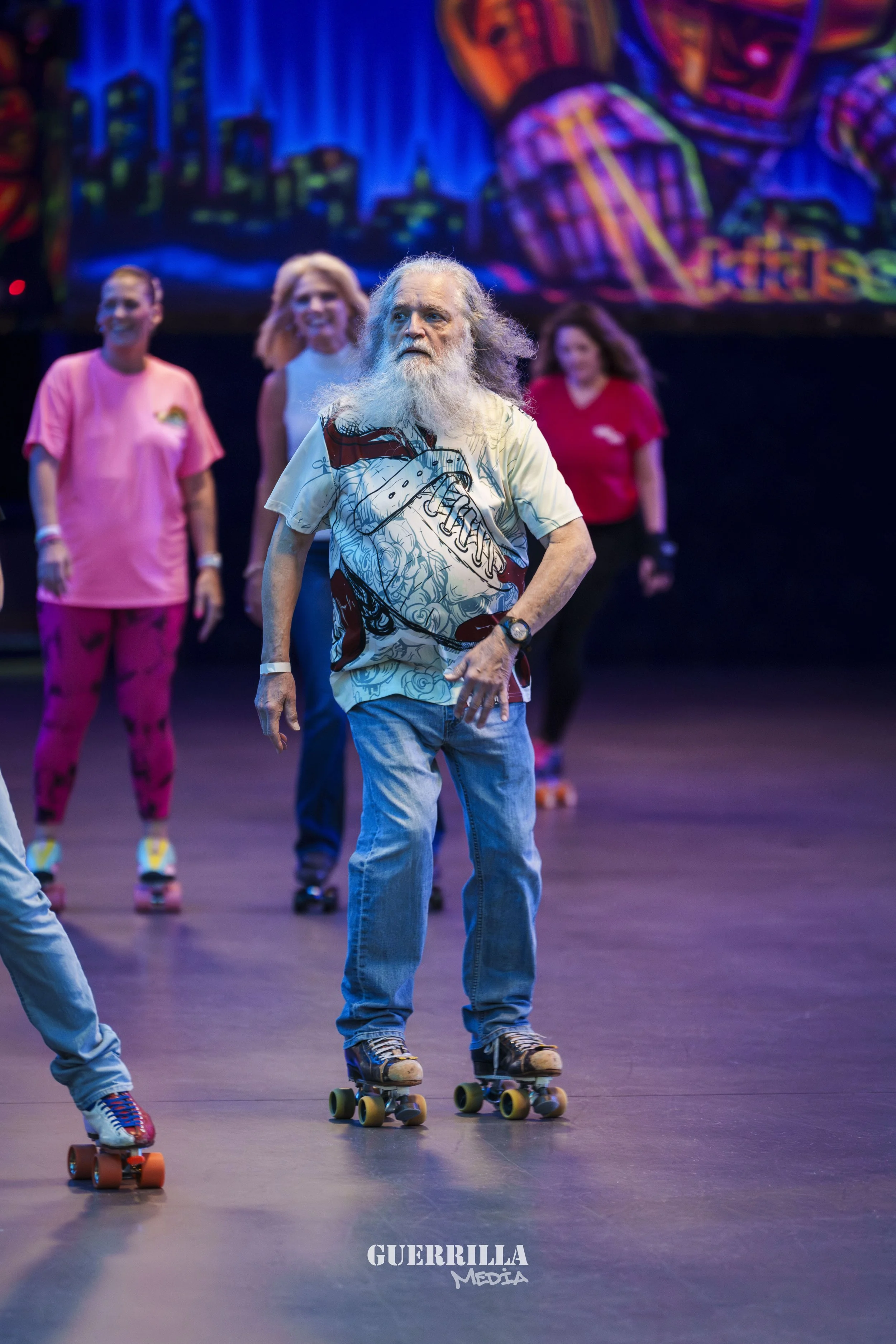 An elderly man with long gray hair and beard roller skating in a group, wearing a colorful shirt and jeans, with a background of a vibrant painted mural and other people on roller skates.