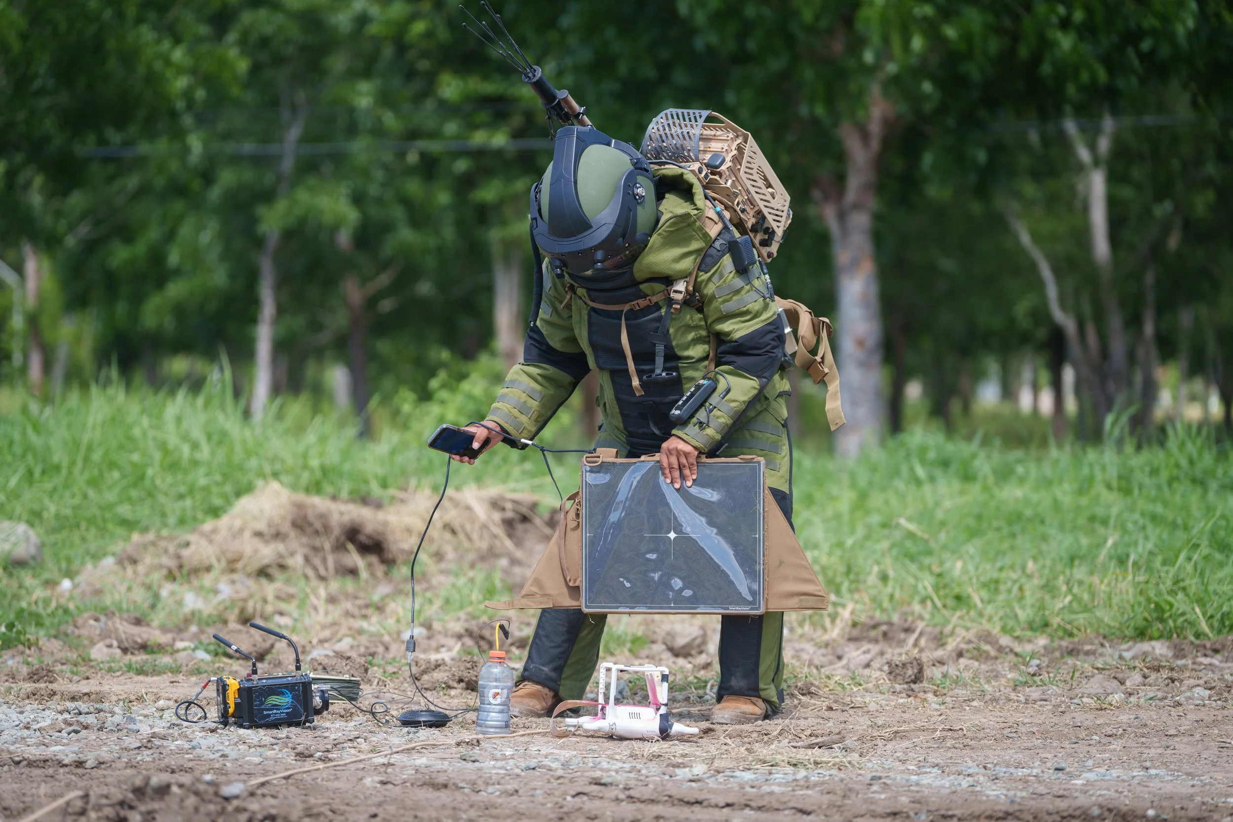 A person in tactical gear outdoors, examining equipment connected to a drone and a small device, with trees in the background.