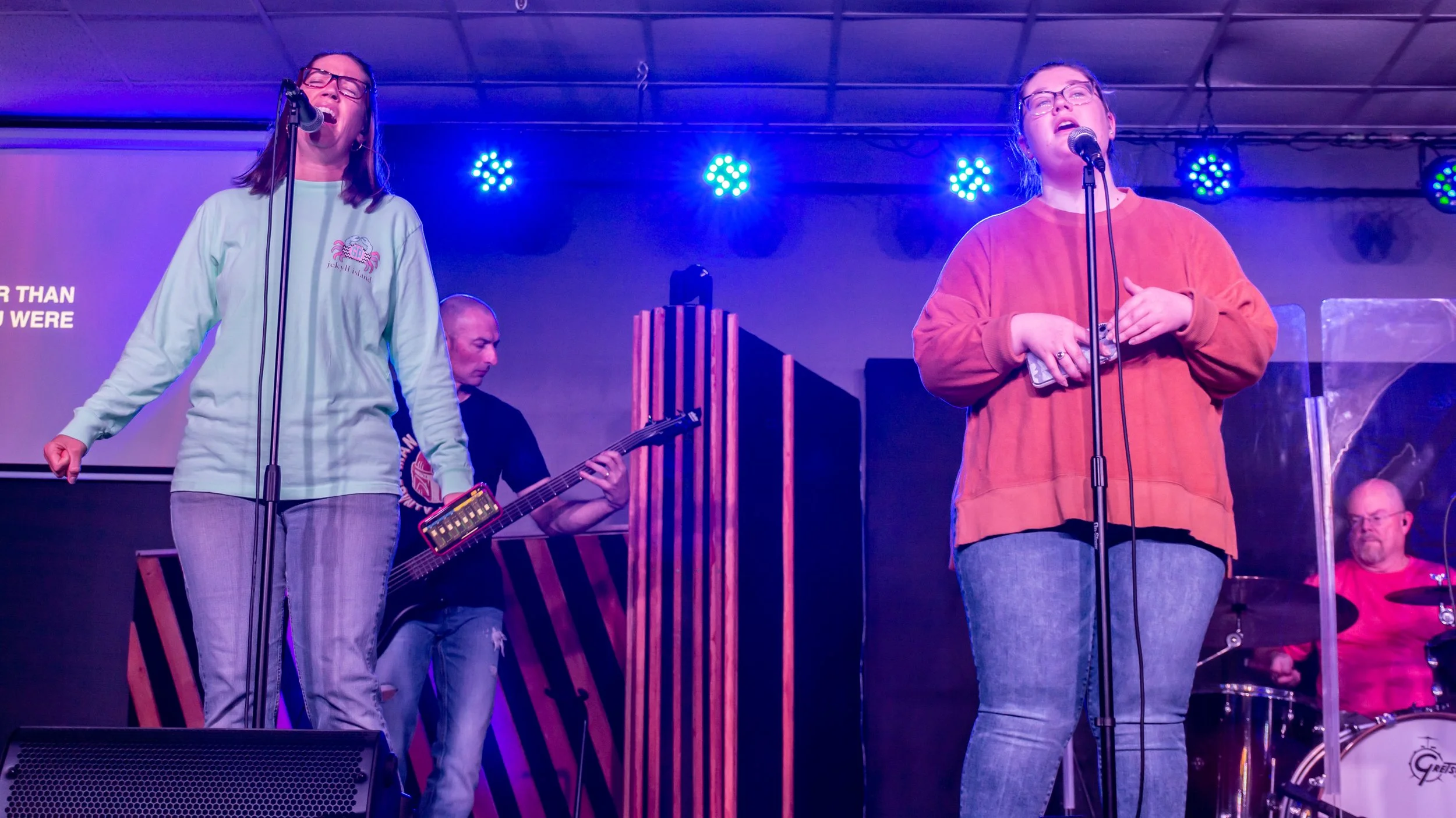 Two women singing into microphones on stage, with a guitarist and drummer in the background, during a live music performance under colorful stage lights.