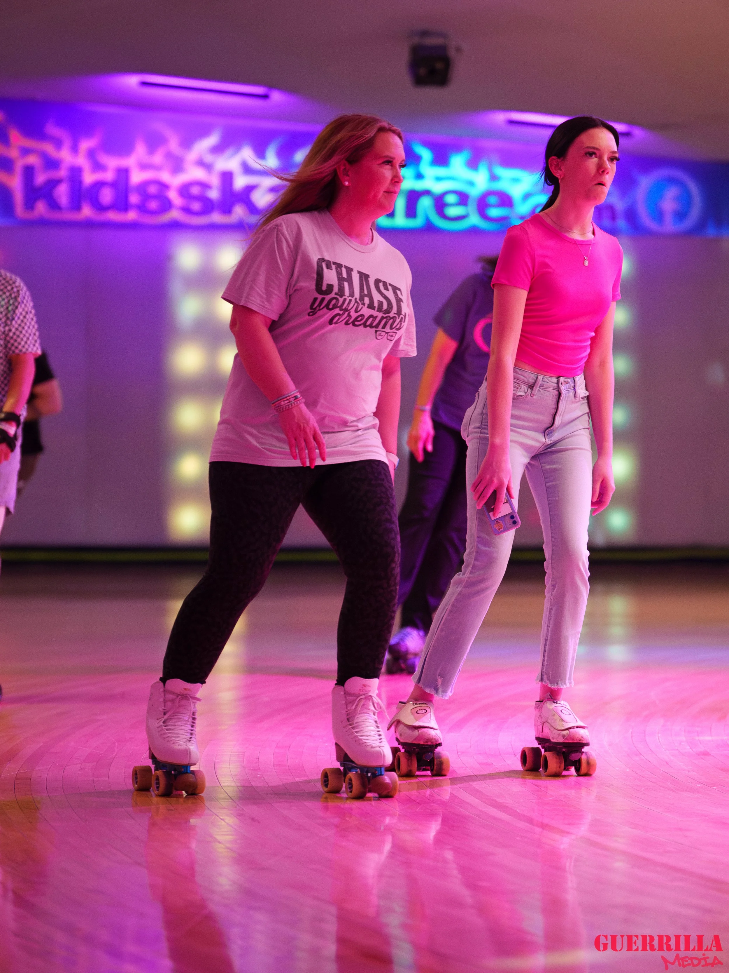 Two young women roller skating in an indoor skating rink with colorful purple and blue neon lights, one wearing a pink shirt and light-colored jeans, the other wearing a white t-shirt with a graphic and dark pants.