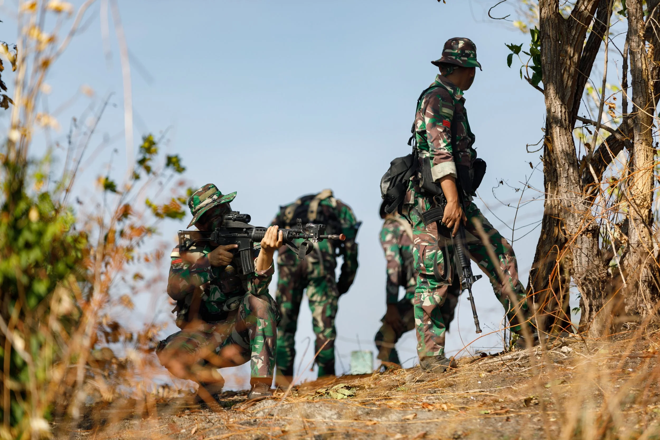 Group of soldiers in camouflage uniforms and gear, armed with rifles, walking through a dry, wooded outdoor area.
