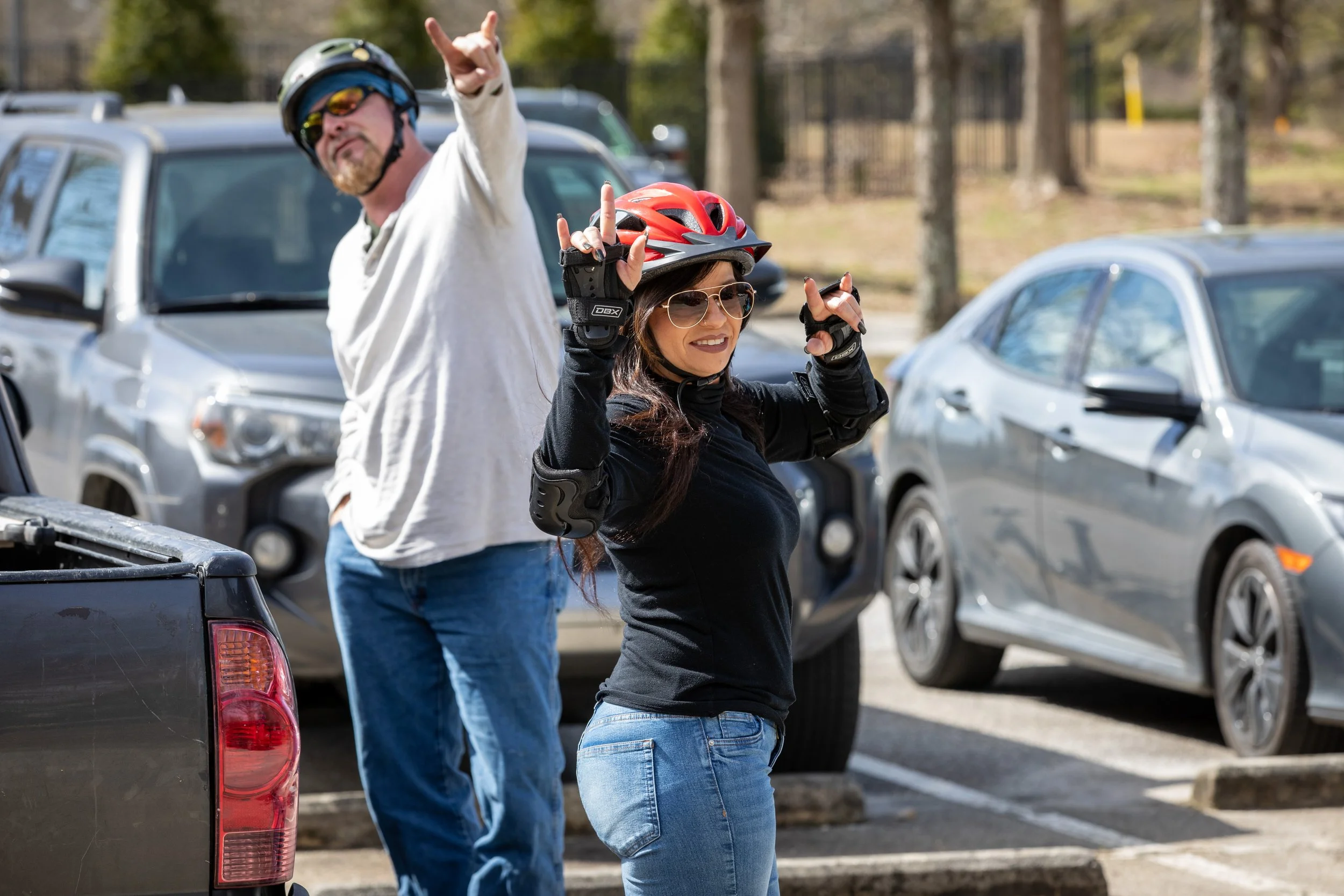 A woman and man in a parking lot, both wearing helmets and protective gear. The woman is also wearing sunglasses and making peace signs with both hands, smiling at the camera. The man is pointing with his right hand, standing behind her, and smiling.