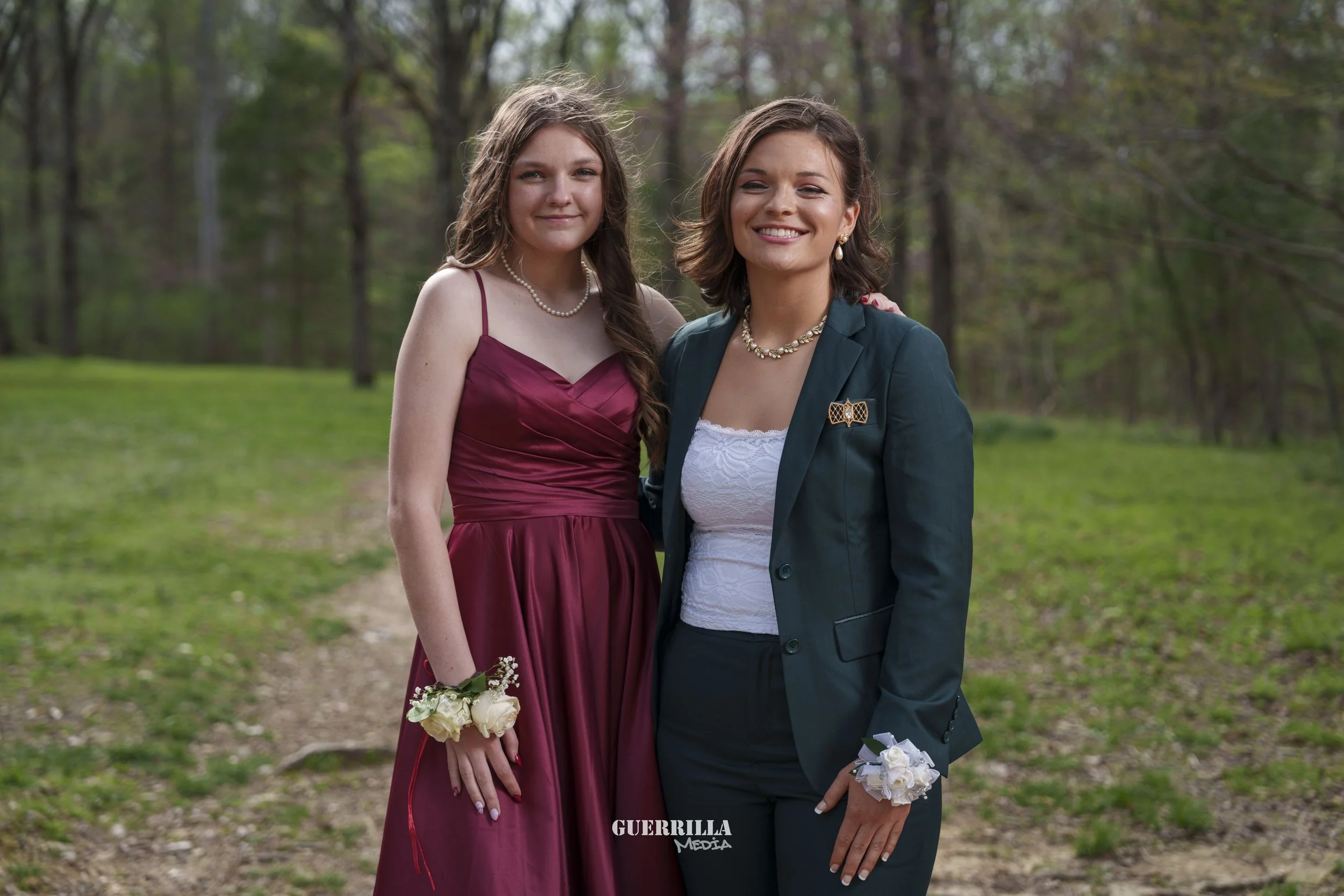 Two young women stand outdoors on a grassy path with trees in the background. One wears a maroon dress with a corsage and pearl necklace, and the other wears a dark blazer with a white top, a brooch, and a wrist corsage.