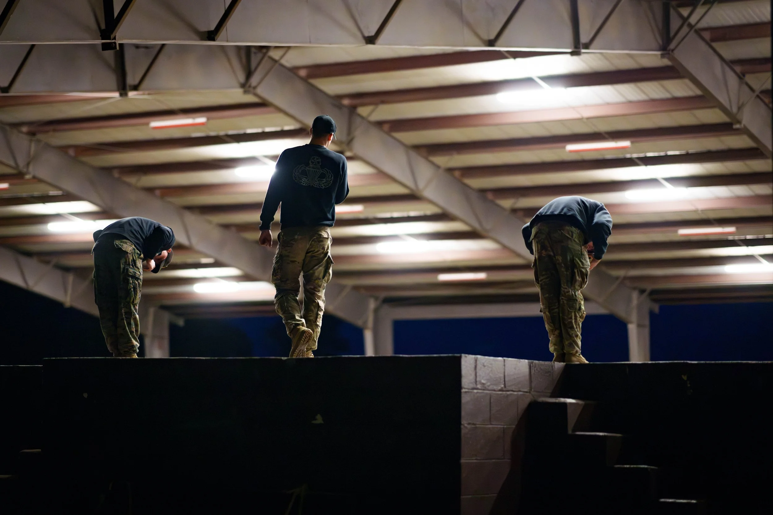 Three individuals, two bent over and one standing, on a raised platform in a large, open structure with a high ceiling, during nighttime.