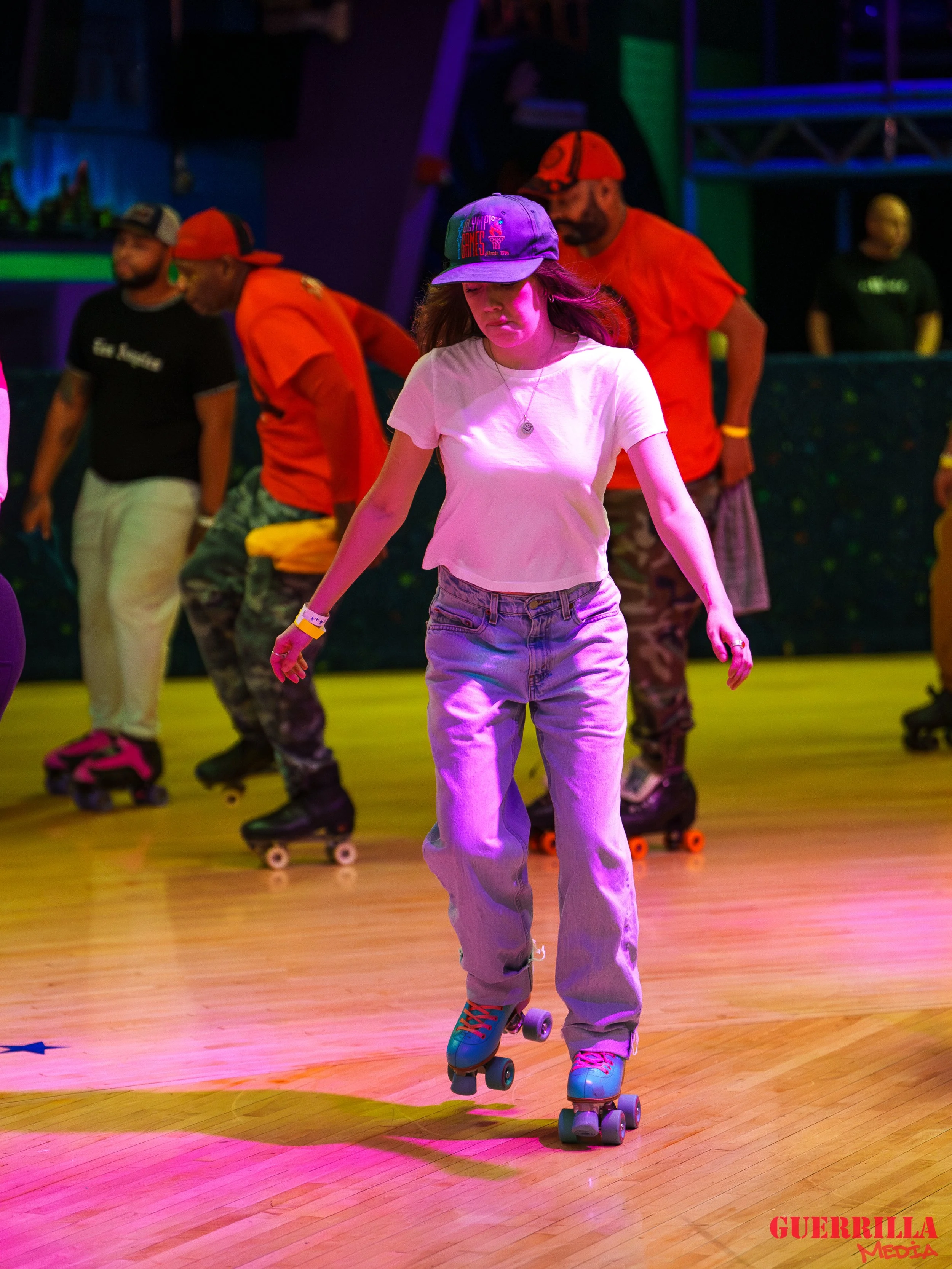 Young woman roller skating on a wooden indoor rink, wearing a white t-shirt, light jeans, a purple cap with colorful writing, and a necklace. In the background, other skaters in casual clothing and a colorful, possibly neon-lit setting, with some wea
