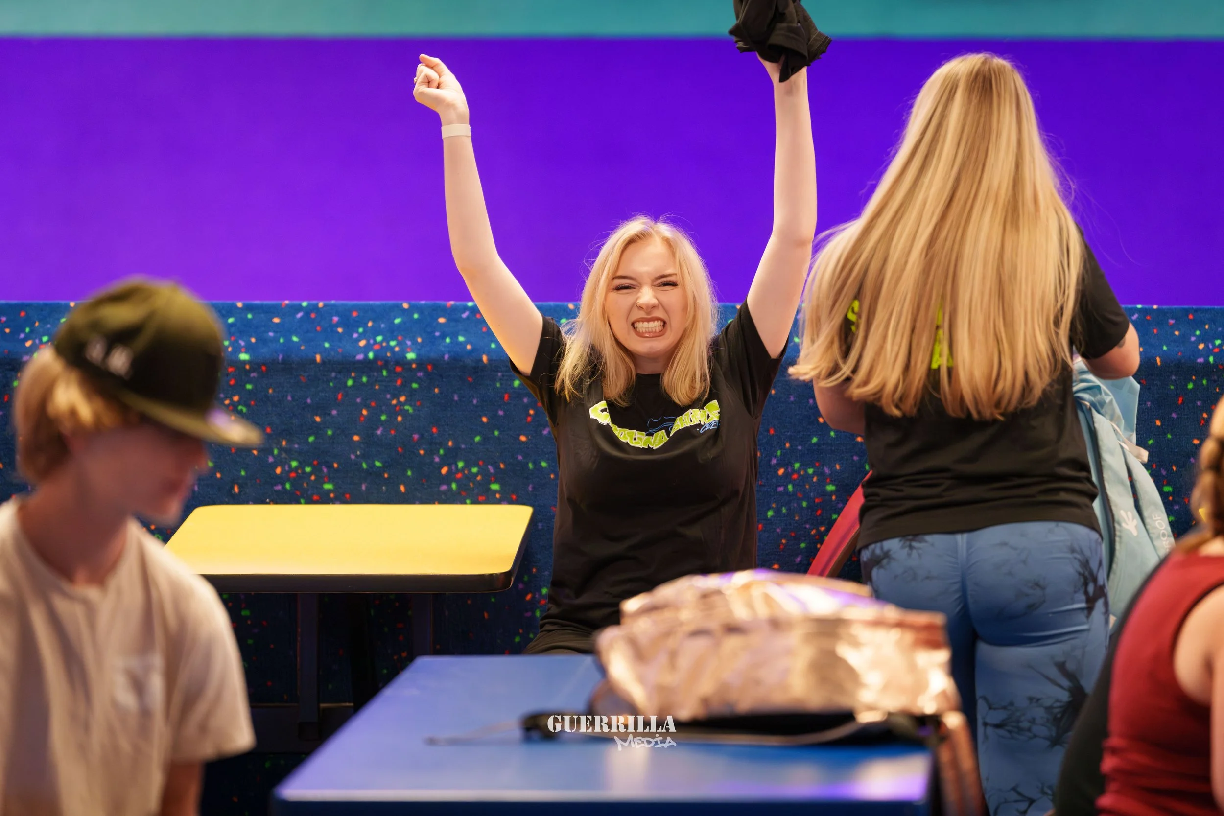 A young woman with blonde hair, wearing a black shirt, is celebrating with her arms raised and a big smile at a social gathering.