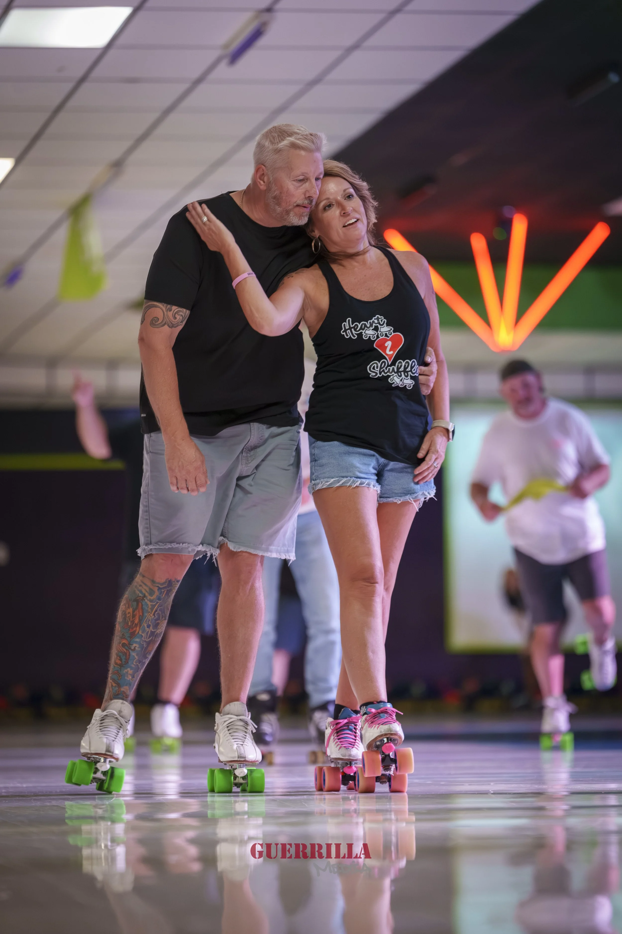 A man and a woman roller skating together indoors. The man has a beard, tattoos on his left arm, and gray hair. The woman has shoulder-length hair and is wearing a black tank top with writing and denim shorts. They are holding each other while skatin