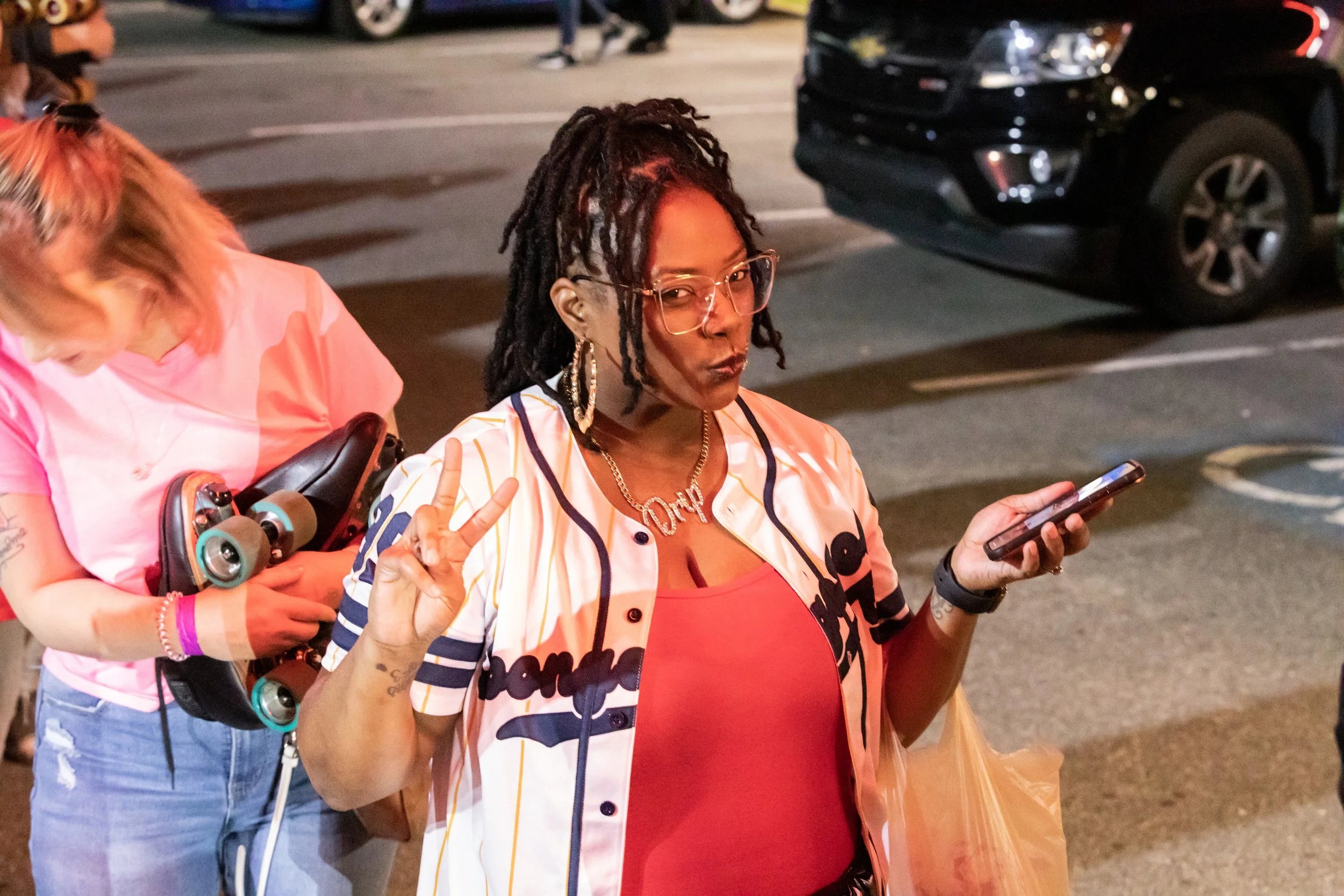 A woman with dreadlocks wearing glasses, earrings, a chain necklace, a red top, and a striped jacket making a peace sign with one hand and holding a phone in the other. She is standing in a parking lot at night.