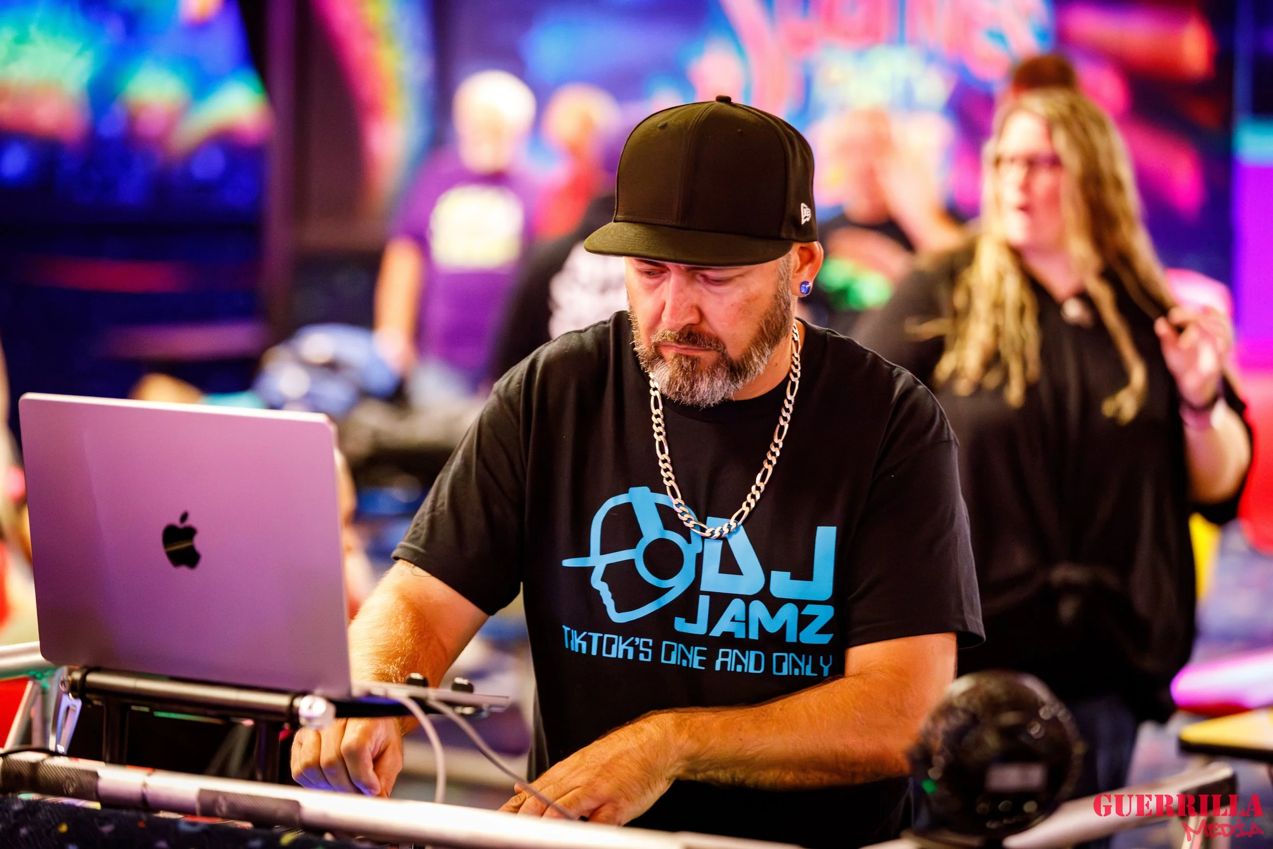 A DJ with a beard, wearing a black baseball cap, black T-shirt with blue text and graphics, and a silver chain, performing with a laptop and DJ equipment at a colorful nightclub or party.