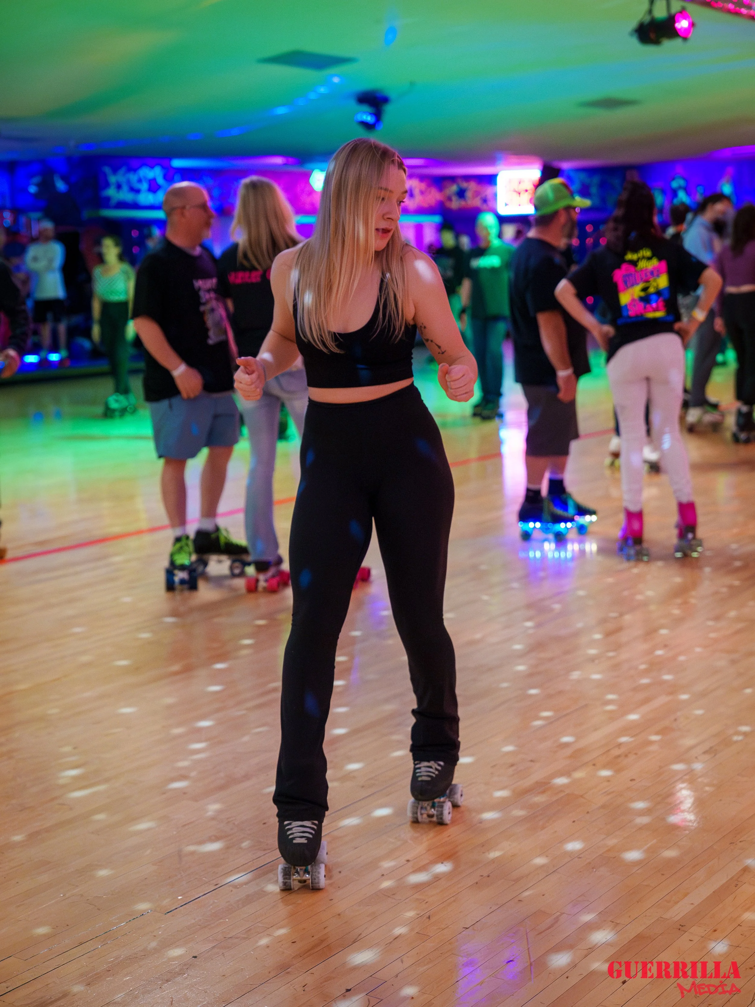 A young woman roller skating in a roller rink with colorful neon lighting, other skaters and a DJ in the background.