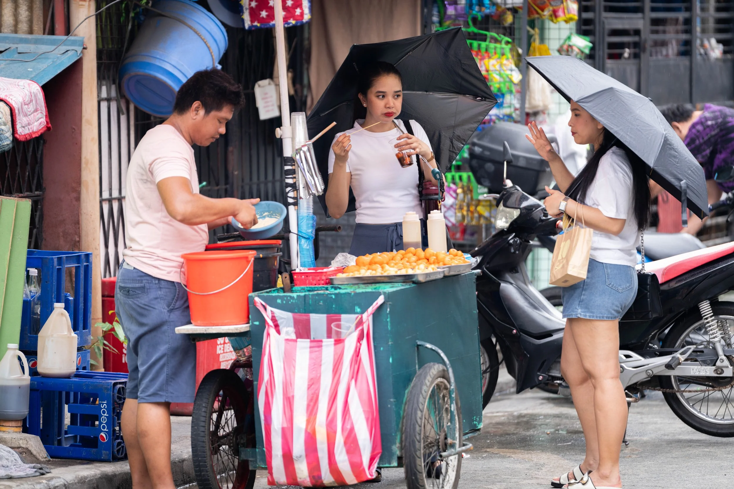 Two women are shopping at a street food stand while a man prepares food. Both women are holding umbrellas, and there are motorcycles parked nearby.