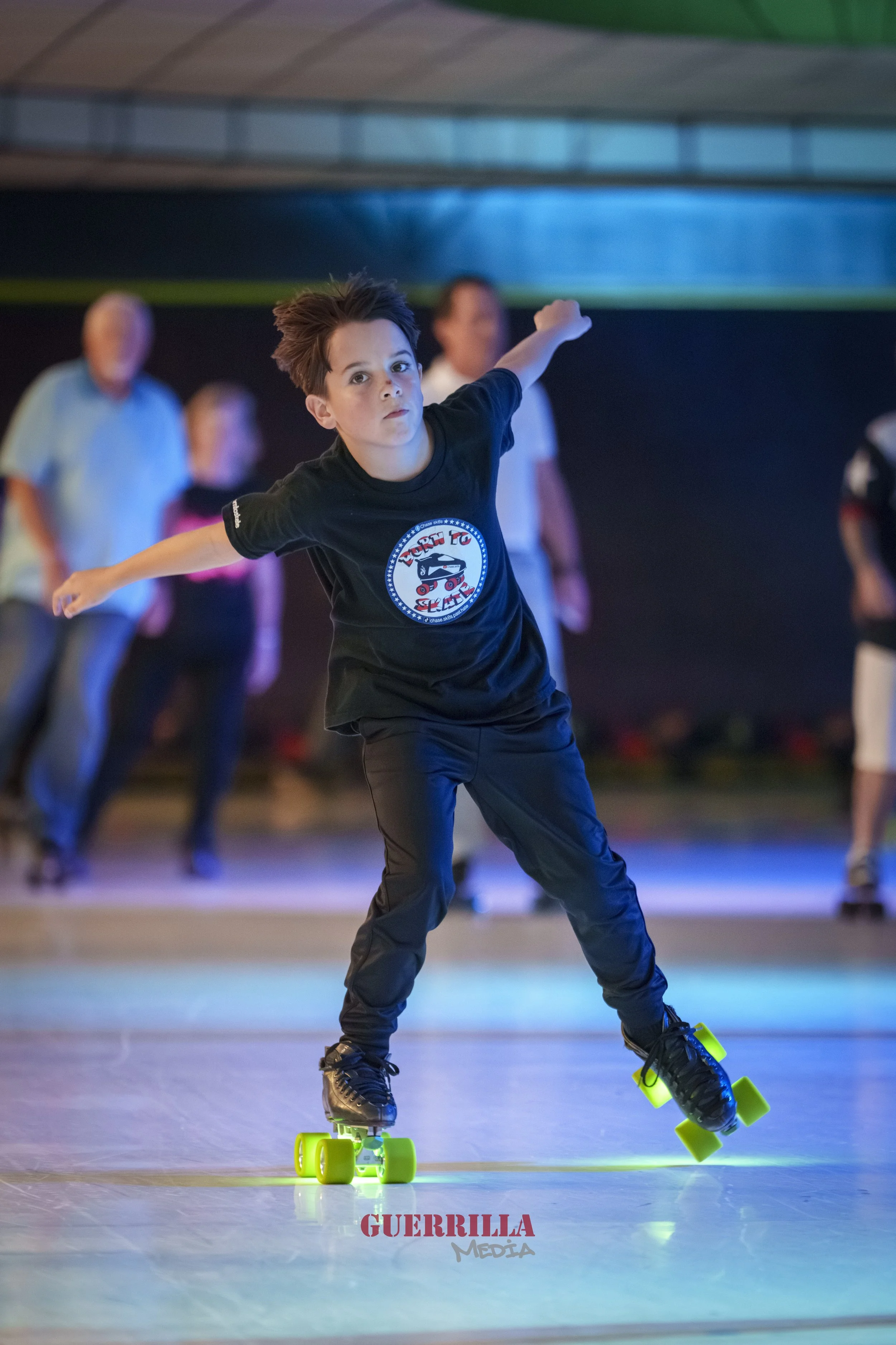 A boy roller skating indoors, surrounded by people, with a focused expression, wearing a black t-shirt with a graphic and black pants.