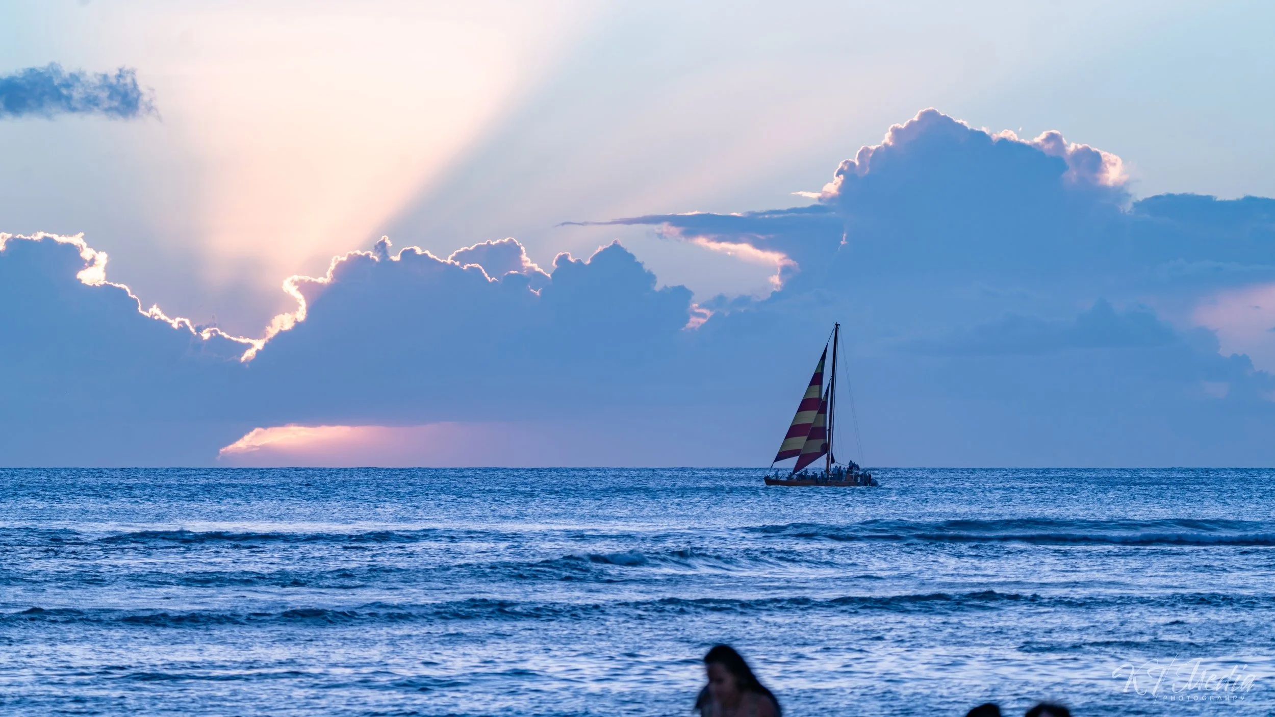 Sunset over the ocean with a sailboat in the distance and a woman in the foreground at the beach.