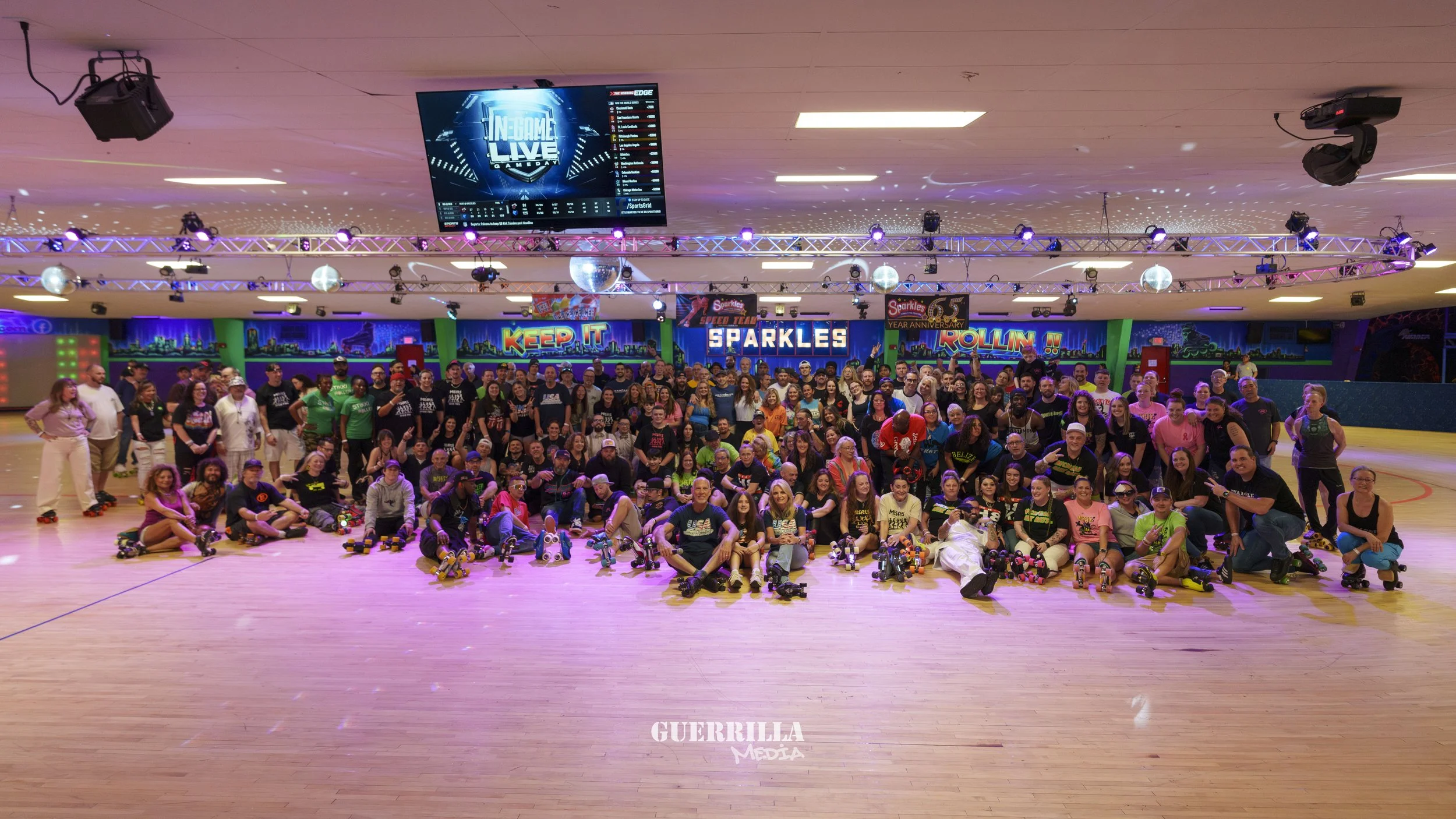 Large group of people at a roller skating rink, some standing and some sitting on the floor with roller skates, colorful lighting, and LED signs in the background.