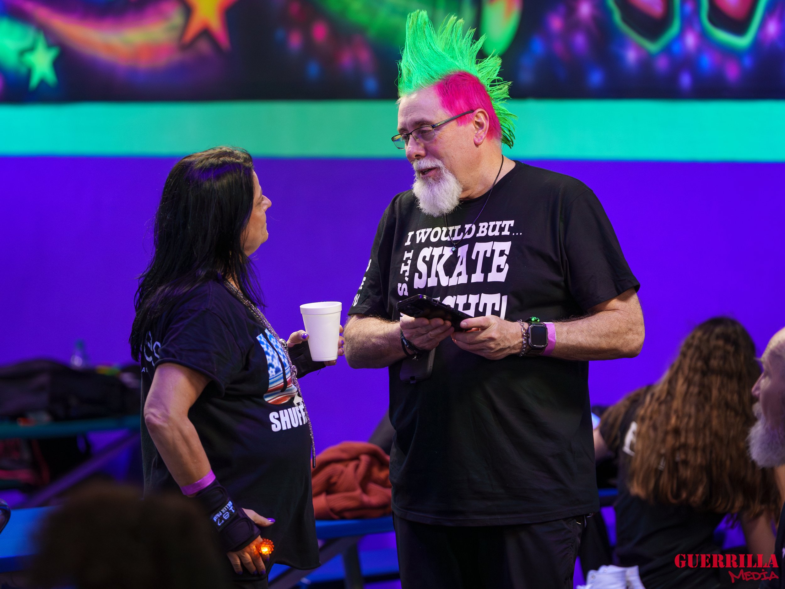 A man with brightly colored green and pink mohawk hairstyle, glasses, gray beard, and black t-shirt with white text, talking to a woman with black hair and a black shirt, holding a white cup inside a colorful, neon-lit room with people in the backgro