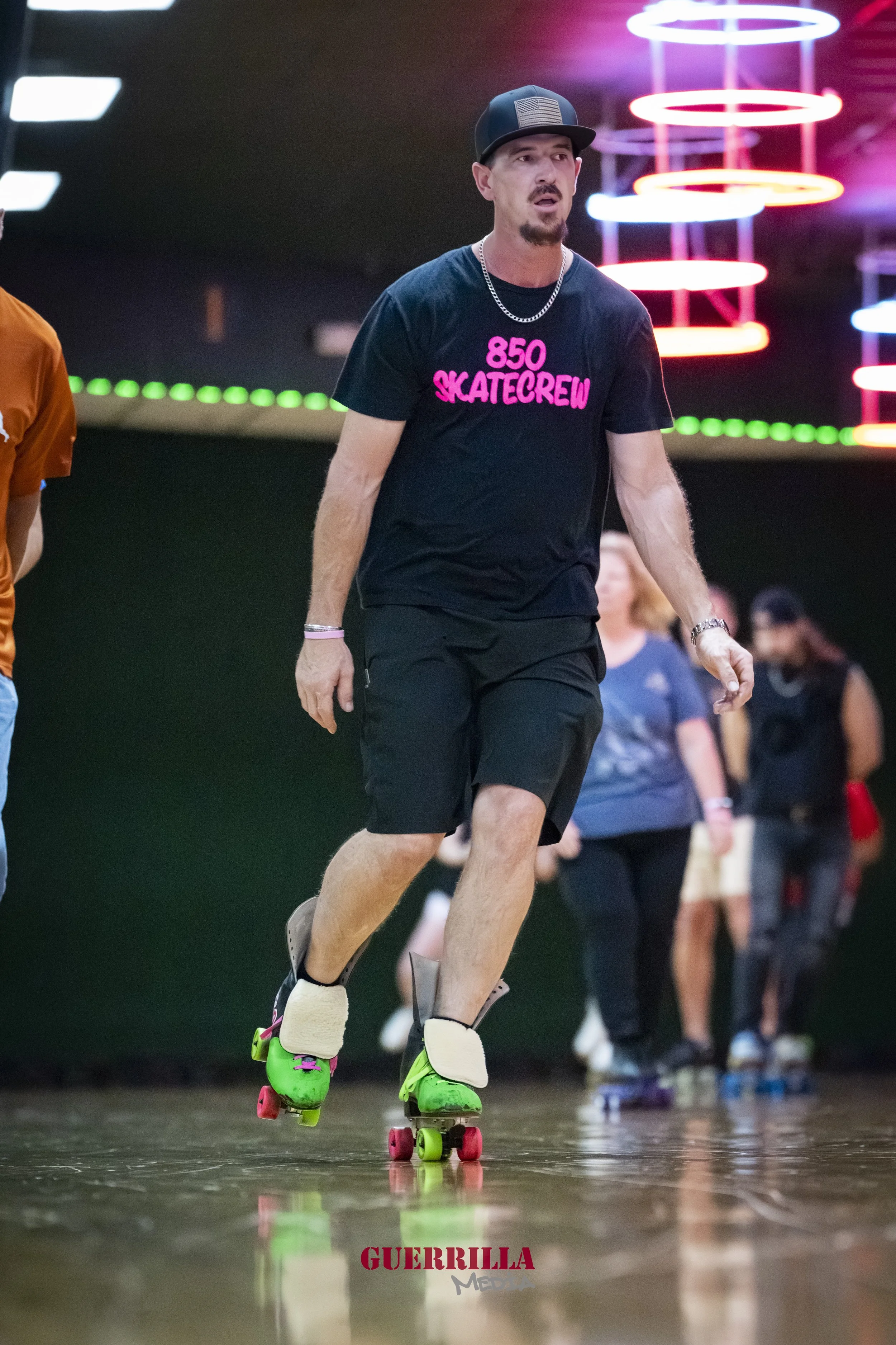A man rollerblading indoors, wearing a black t-shirt with pink text that says '850 Skatecrew', black shorts, and a black baseball cap, with neon lighting in the background.