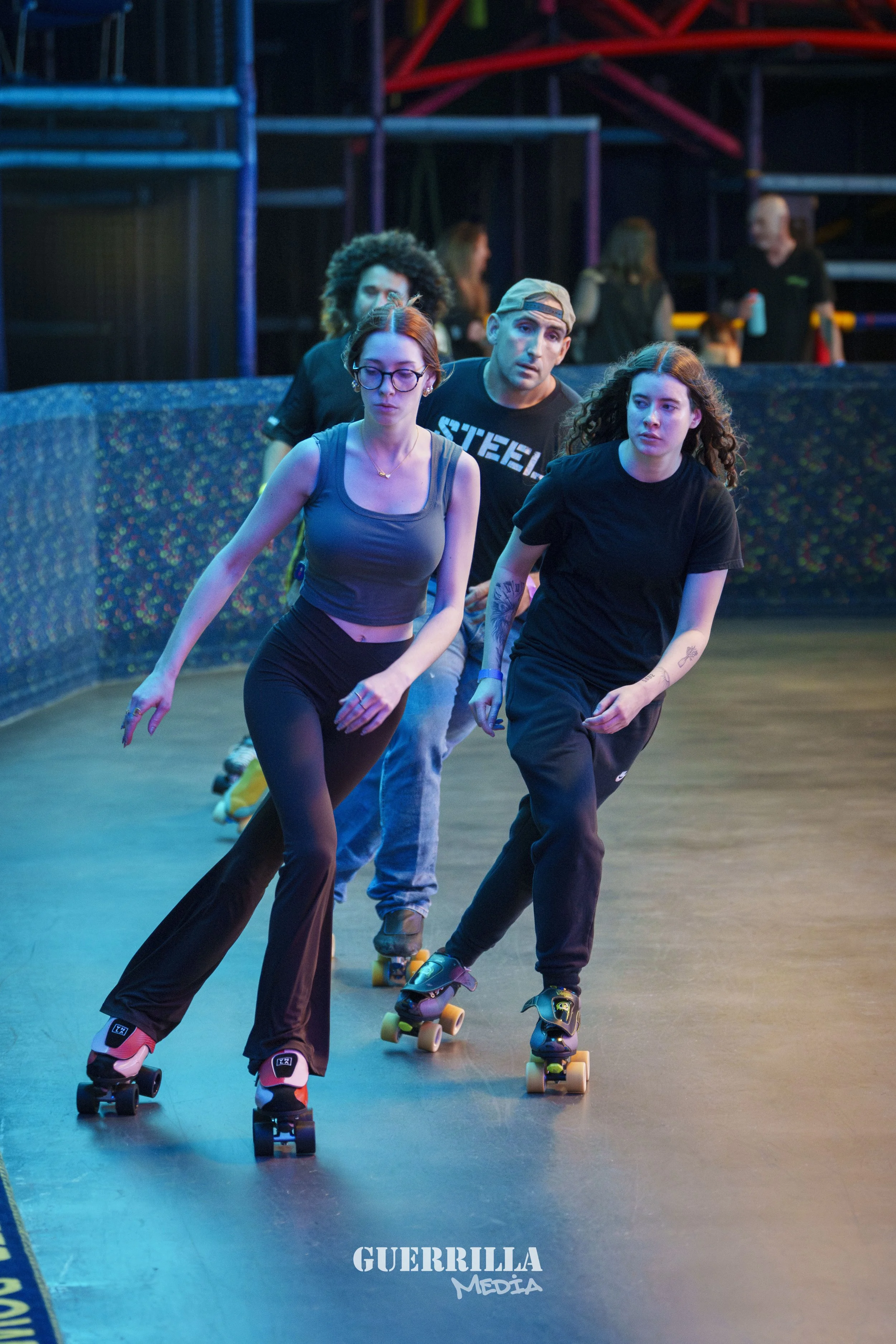 Three people skating in an indoor roller rink, with others in the background, and a 'GUERRILLA MEDIA' sign on the floor.