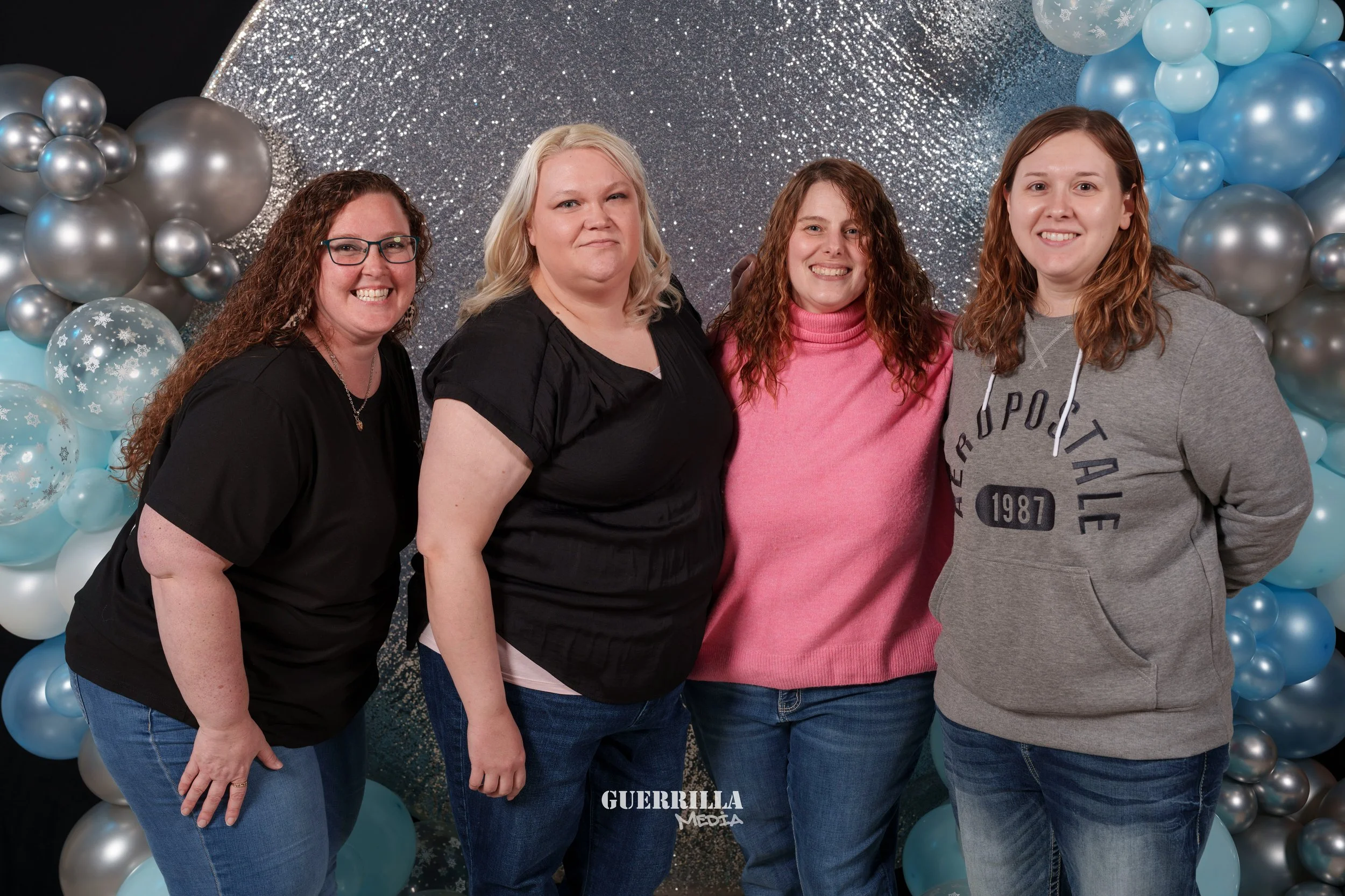 Four women standing in front of a glittery silver and blue balloon backdrop, smiling at the camera.