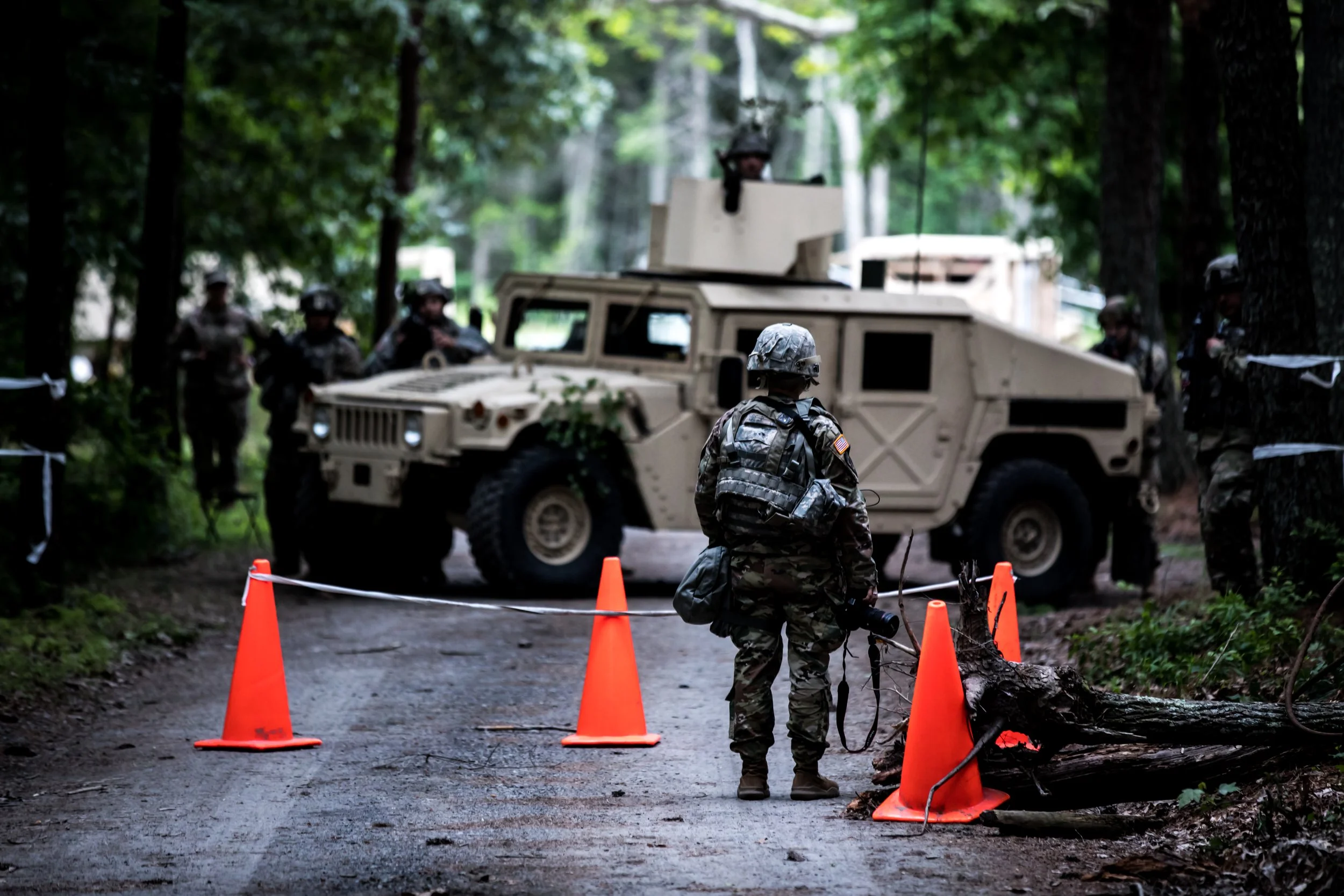 Military personnel and a military vehicle in a wooded area, with orange traffic cones and barriers surrounding a fallen tree.