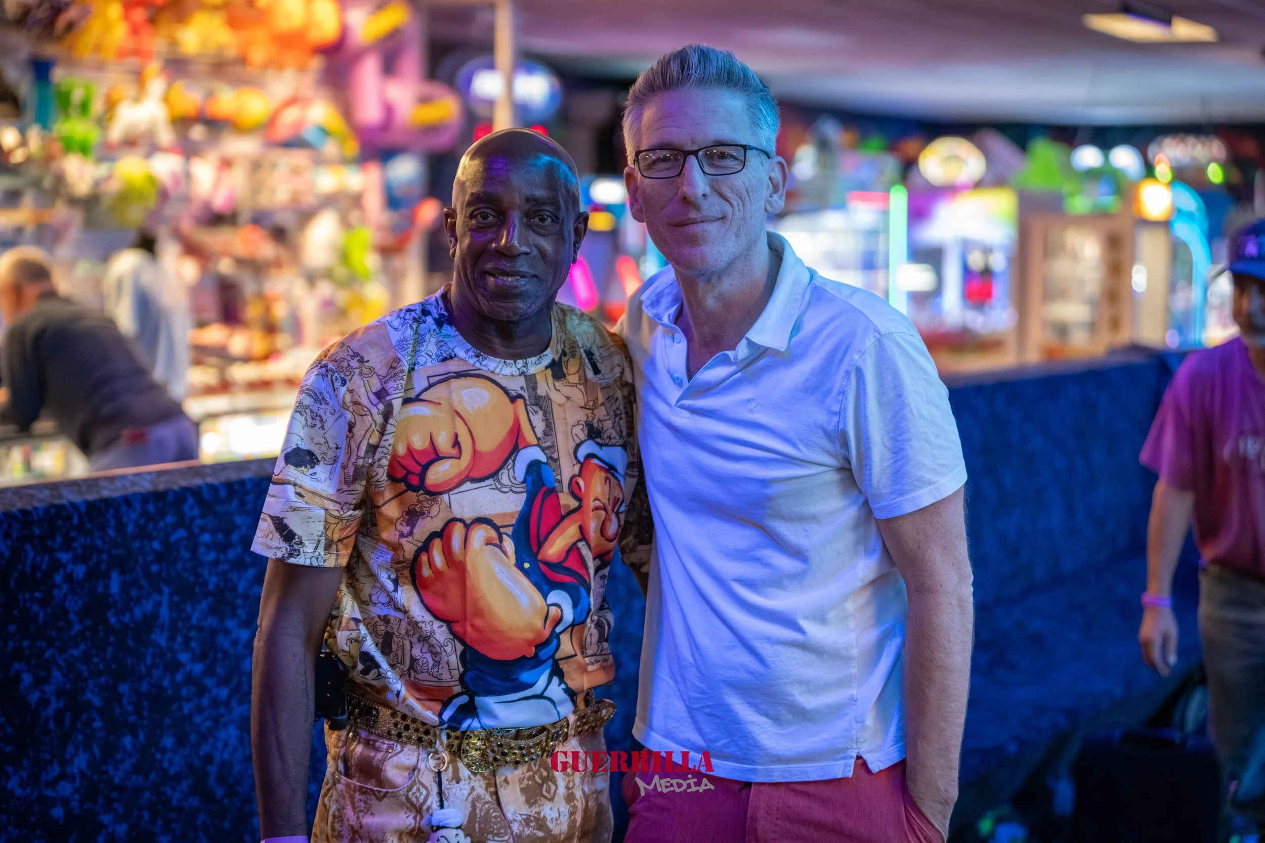 Two men standing close together at a lively fair with colorful lights and game booths in the background.