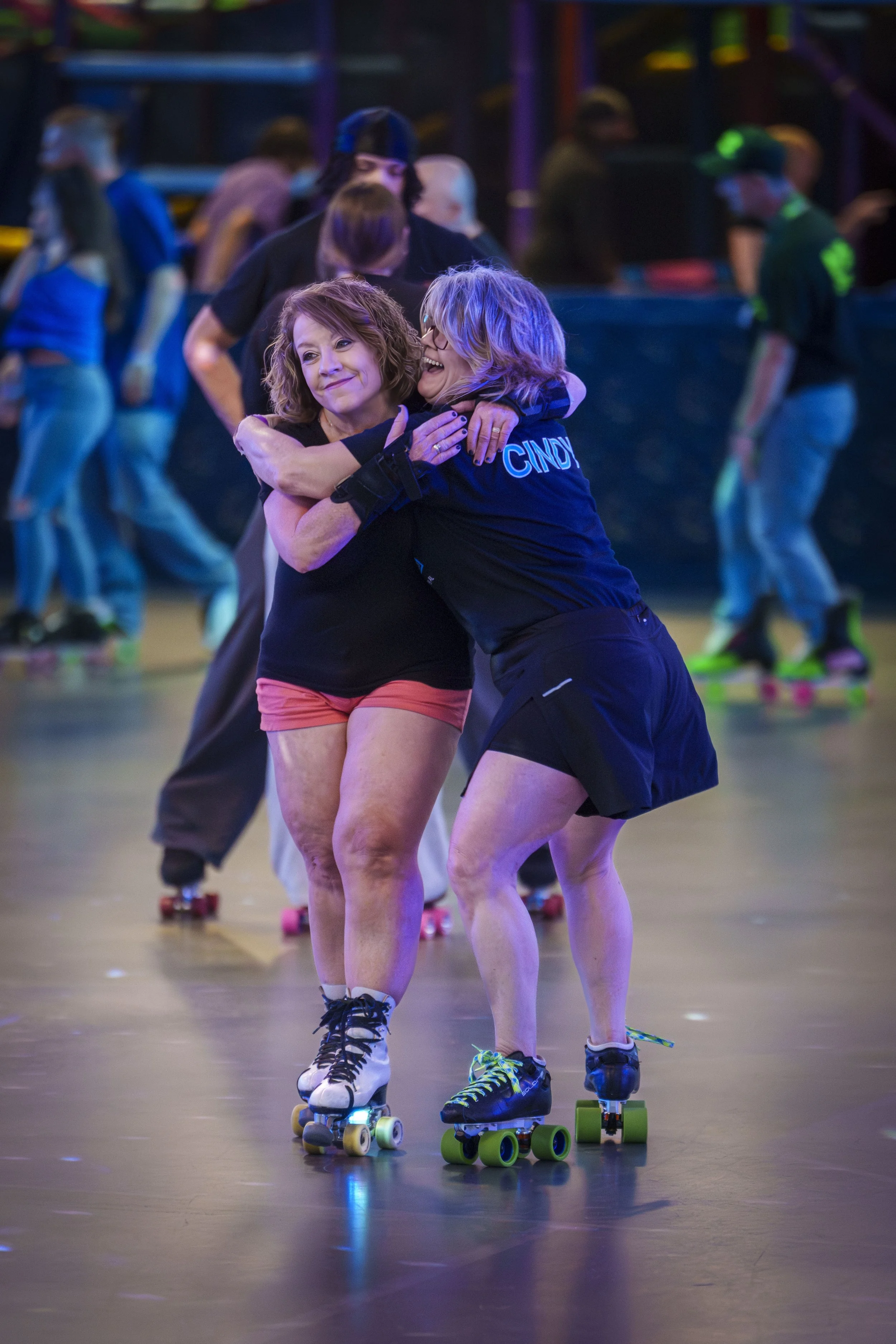 Two women hugging and roller skating in a roller rink, with other skaters in the background.