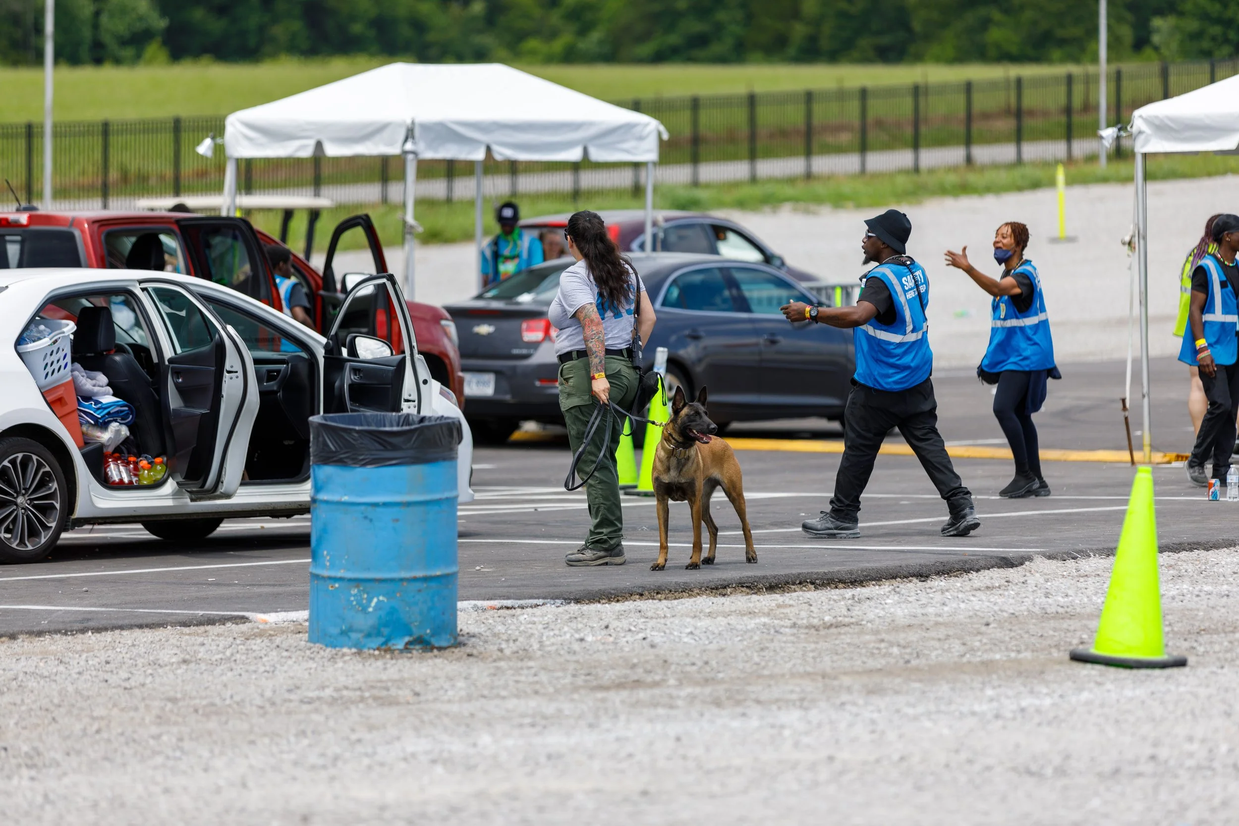 A group of people, including security personnel with a dog, at a parking lot with tents and cars.