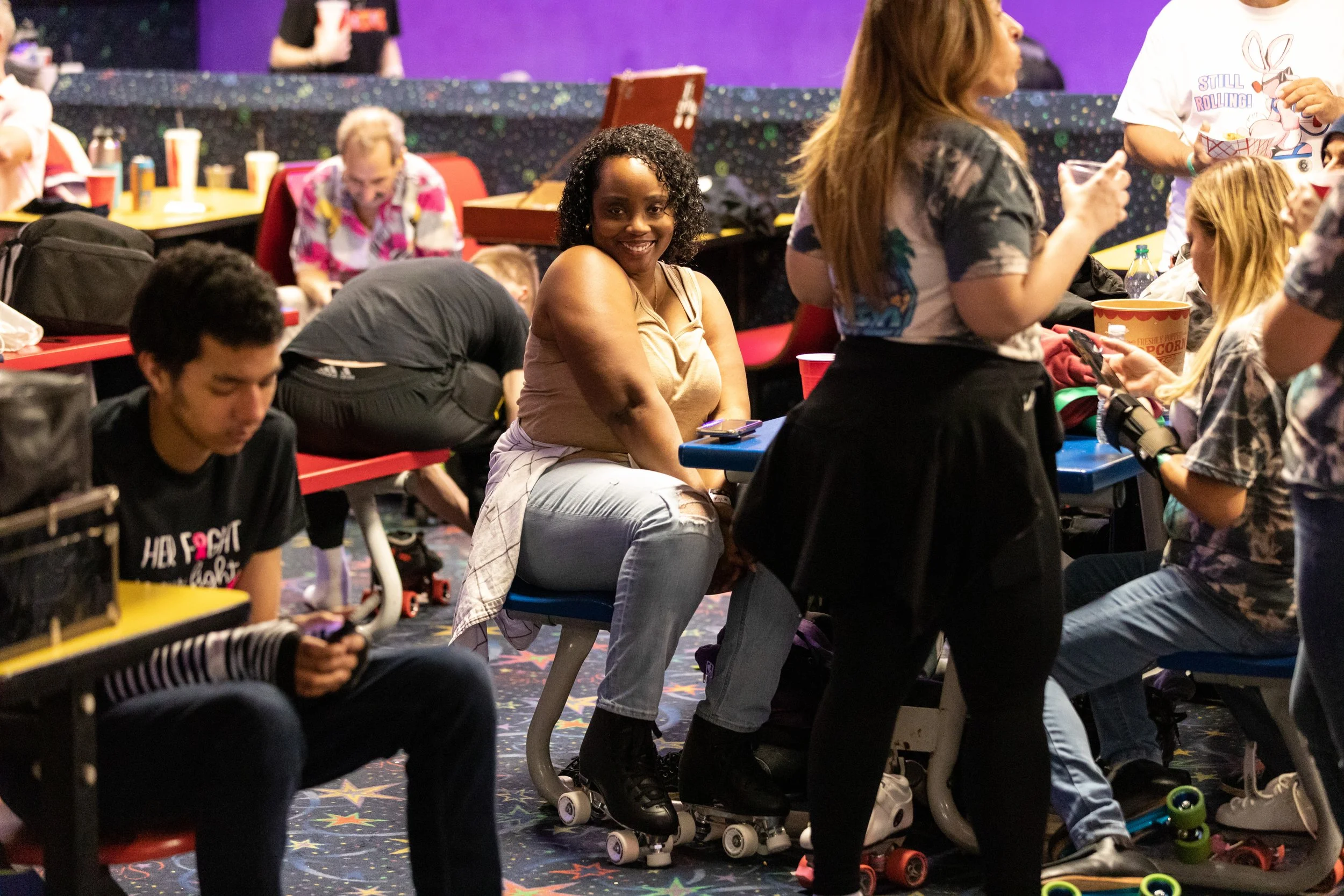 A group of people at a roller skating rink, with some sitting and some standing, engaging with their phones, and a woman in the center smiling at the camera.