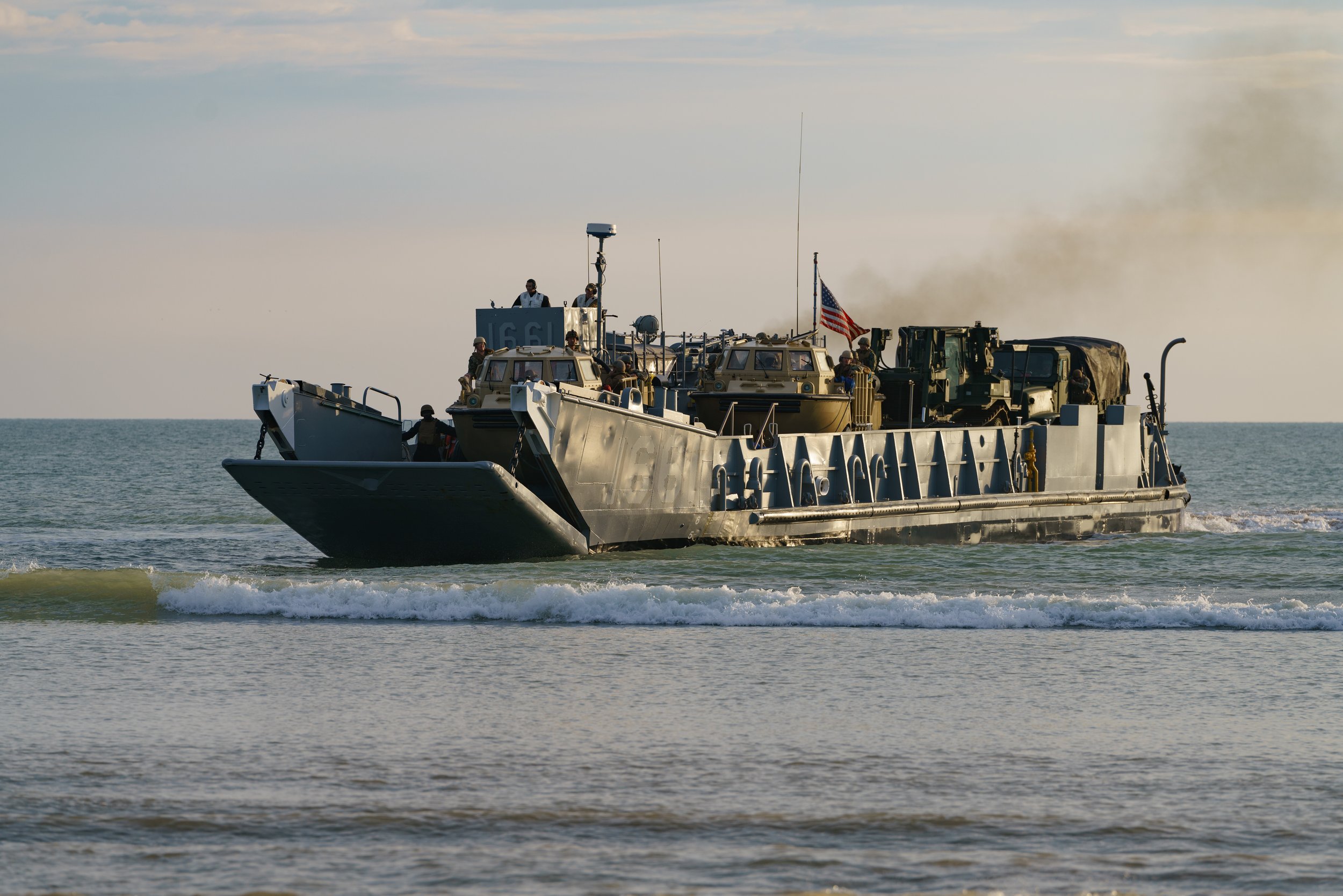Military boat with soldiers and equipment moving through the ocean, with American flag visible.