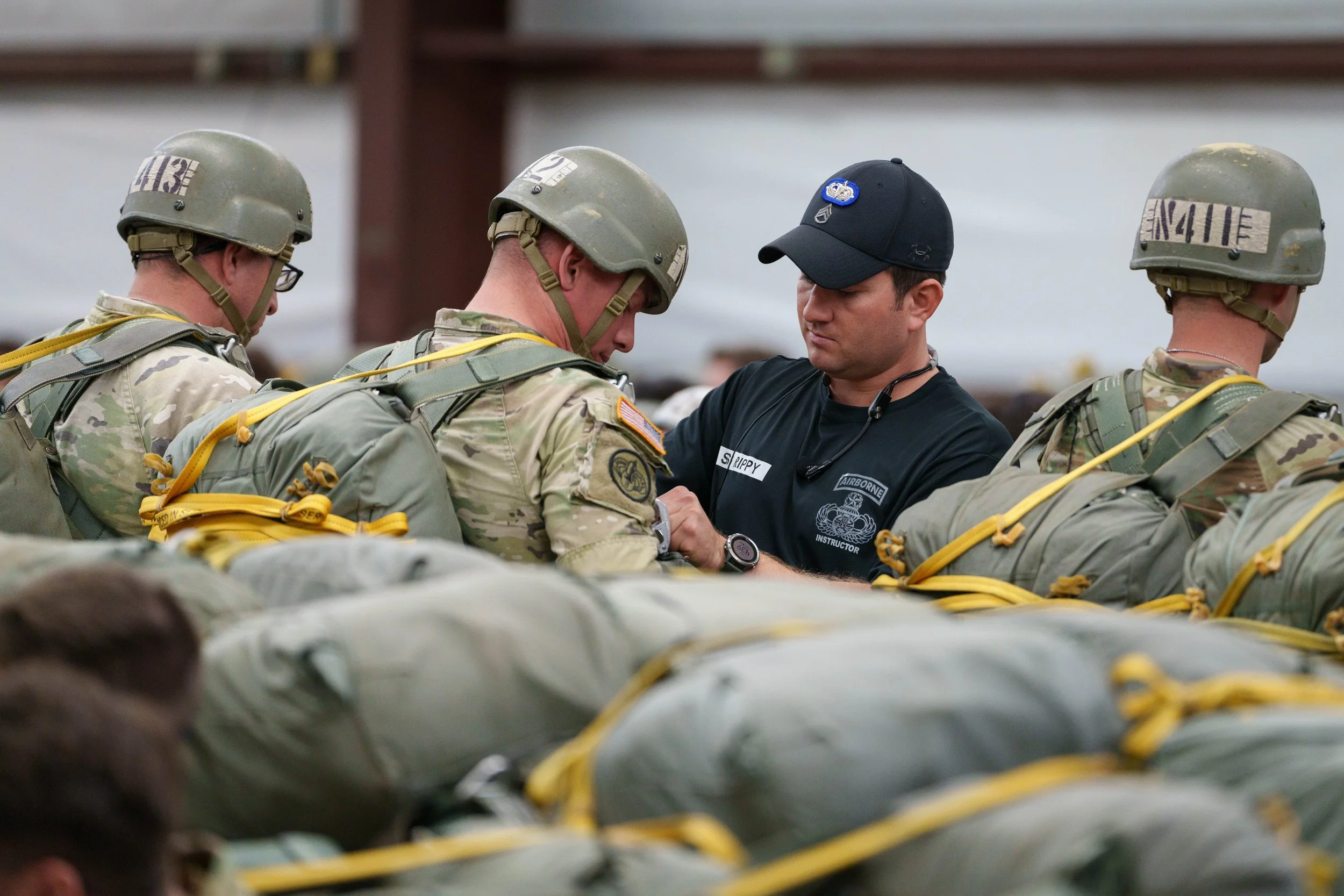 A group of soldiers receiving instruction from a man wearing a black shirt and cap, inside a large indoor space.