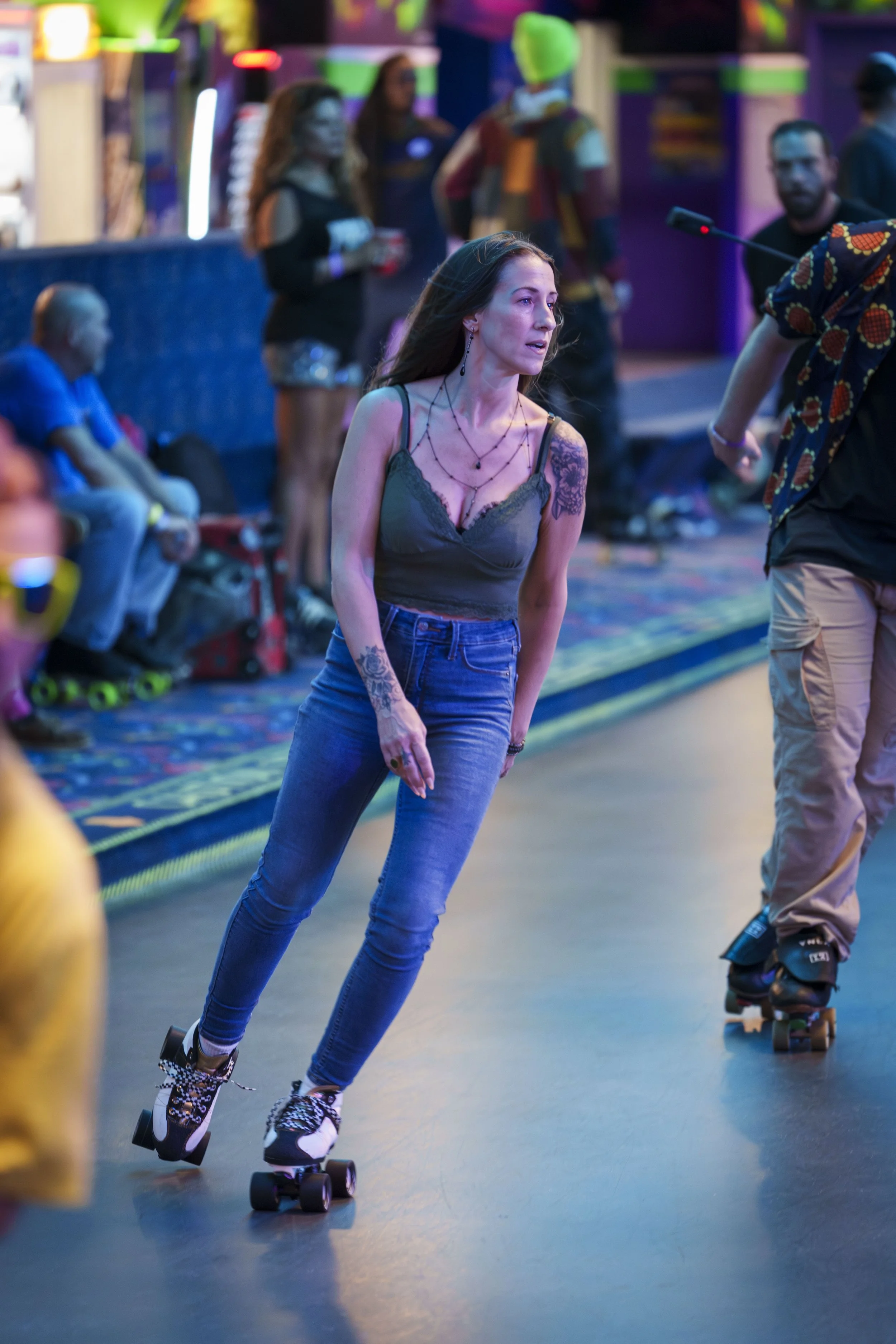 A woman roller skates at an indoor skating rink with colorful neon lights, wearing a green camisole and blue jeans, surrounded by other skaters and spectators.
