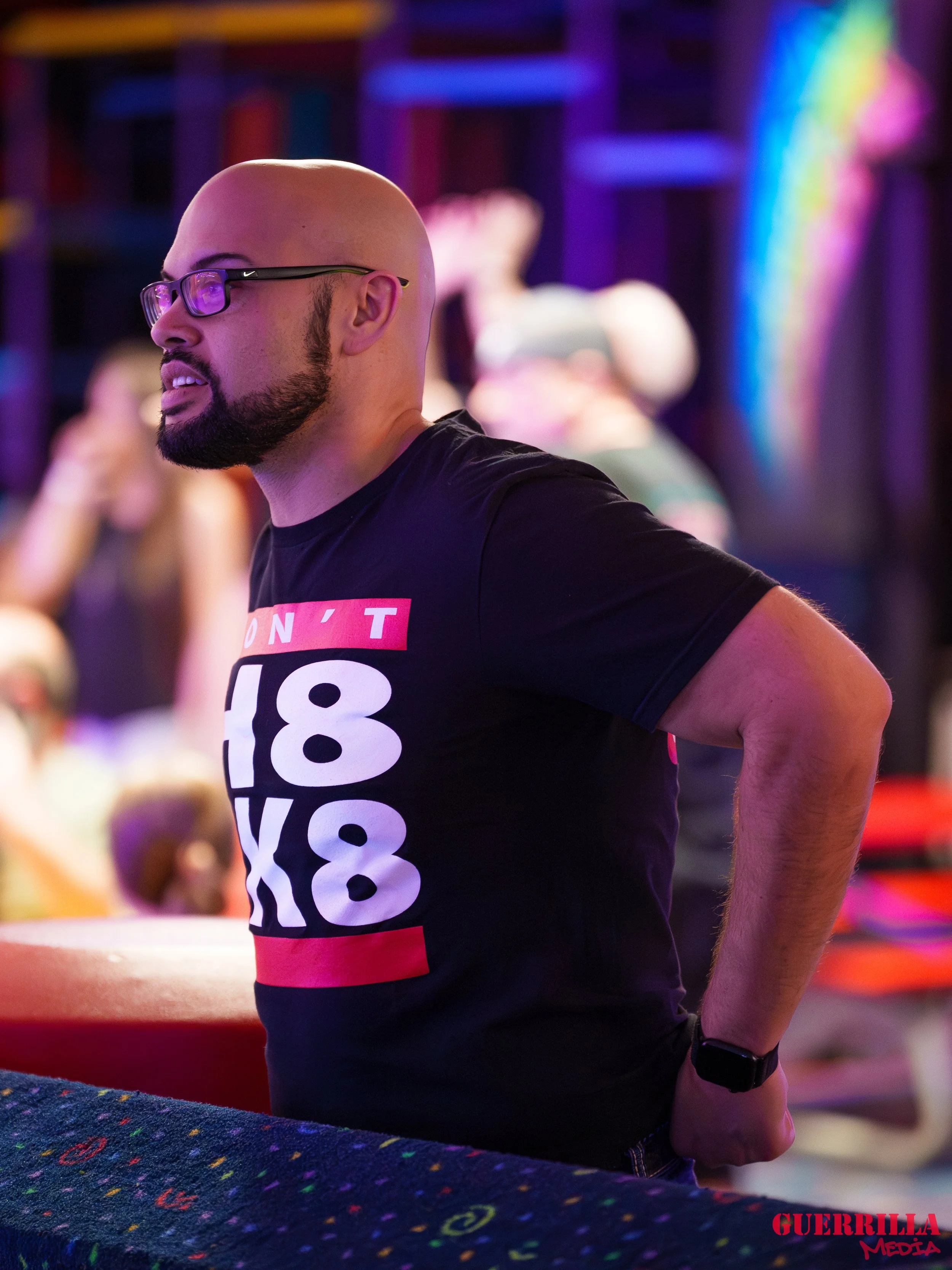 A man with glasses and a beard wearing a black T-shirt with bright pink, white, and red text at a social event with colorful lighting and blurred background of other people.