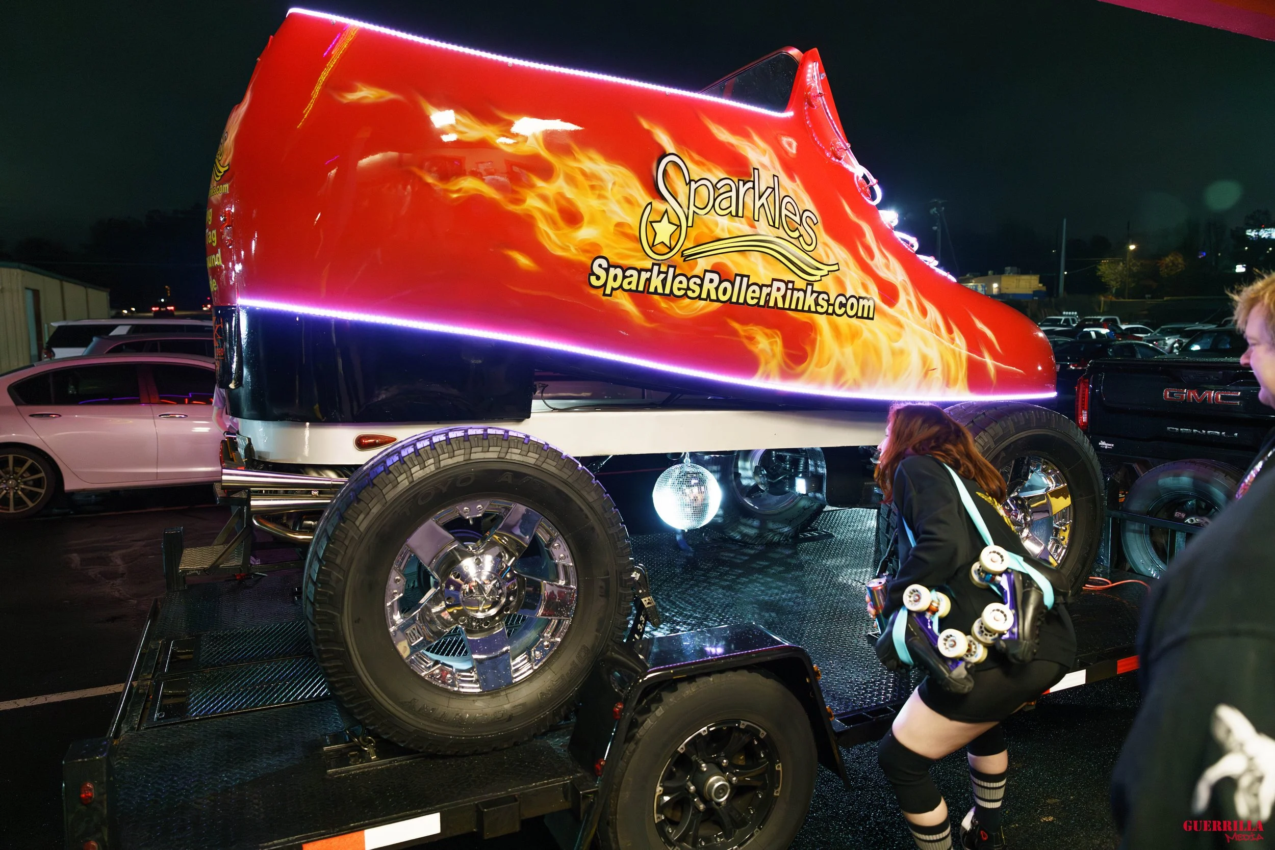 A monster truck with bright red and yellow flame graphics, lit with LED lights, branded 'Sparkles' with the website SparklesRollerRinks.com, parked on a trailer at night, surrounded by people.