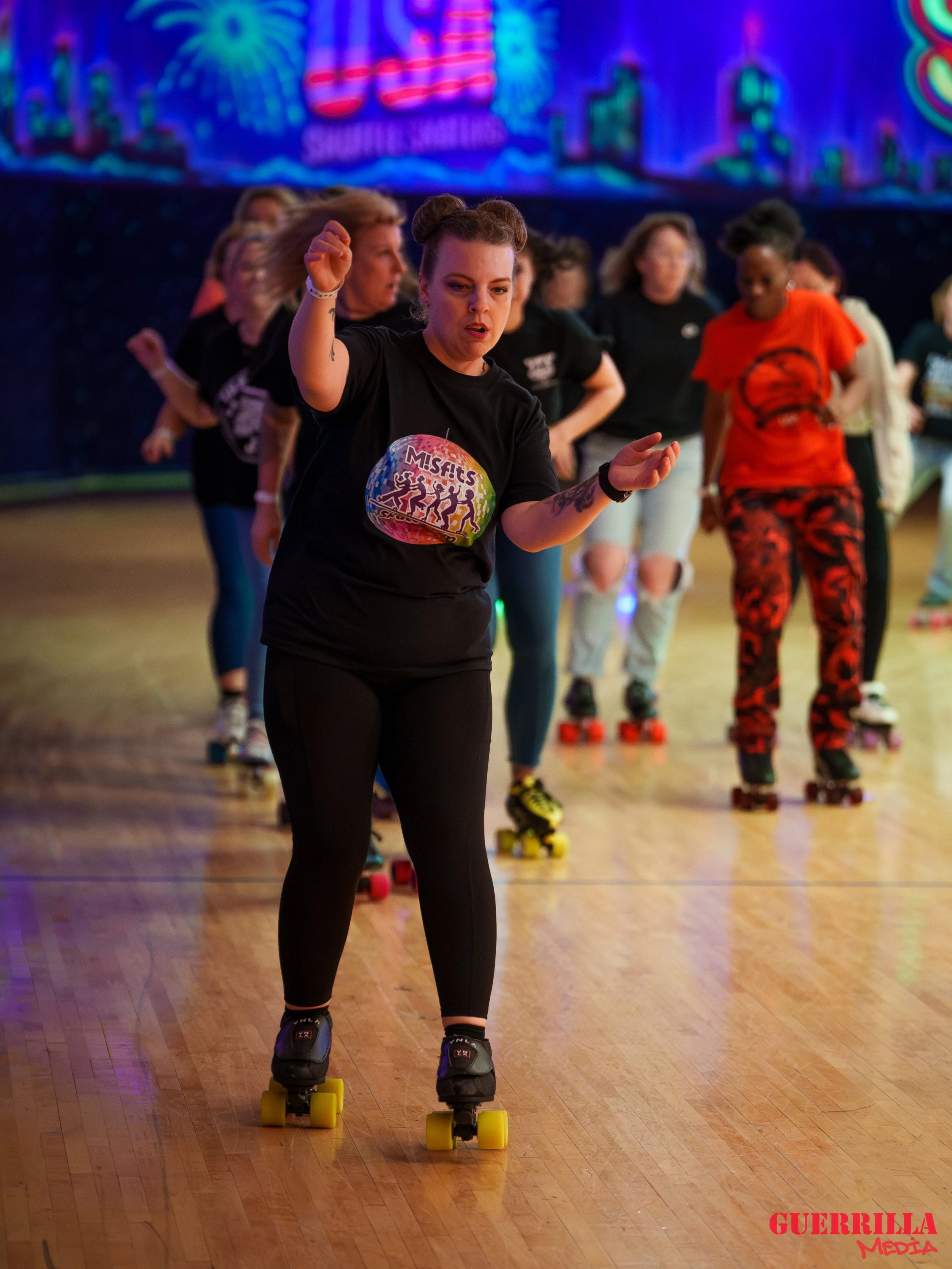 A group of women roller skating indoors on a wooden floor, with a colorful, neon-lit cityscape background. The woman in front is wearing a black Misfits T-shirt and black leggings, while others wear various casual outfits. The scene appears lively an