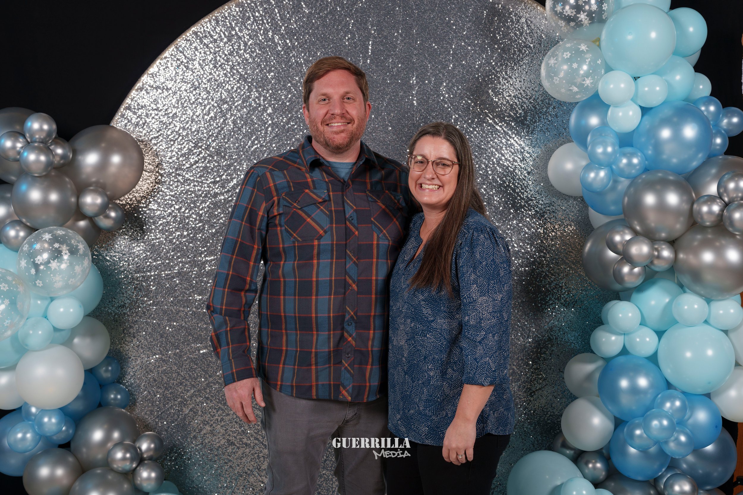 A happy couple stands close together in front of a glittery silver backdrop with balloons in shades of blue, white, and silver arranged on either side.