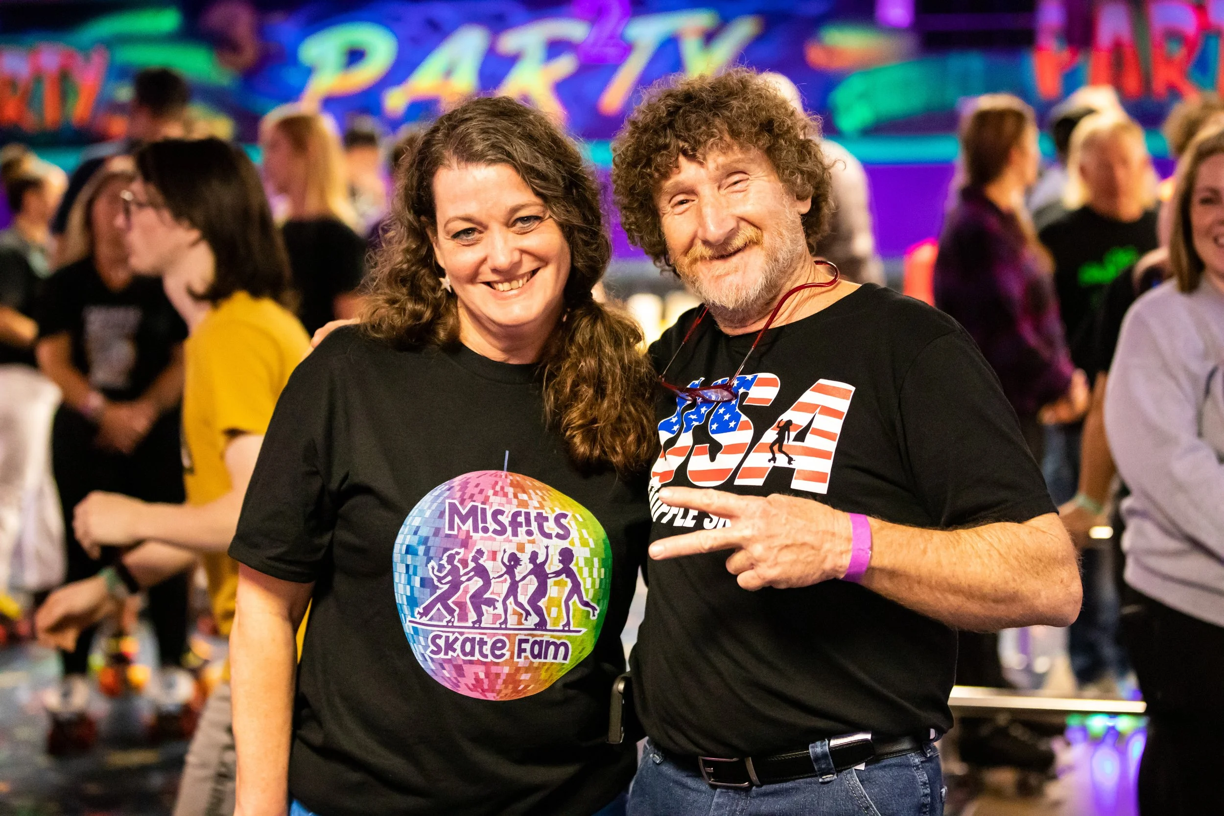 Two smiling people at a roller skating event, one woman wearing a black shirt with a colorful disco ball graphic and the text 'MISFITS Skate Fam,' and one man wearing a black shirt with a patriotic design, making a peace sign. There are other people 