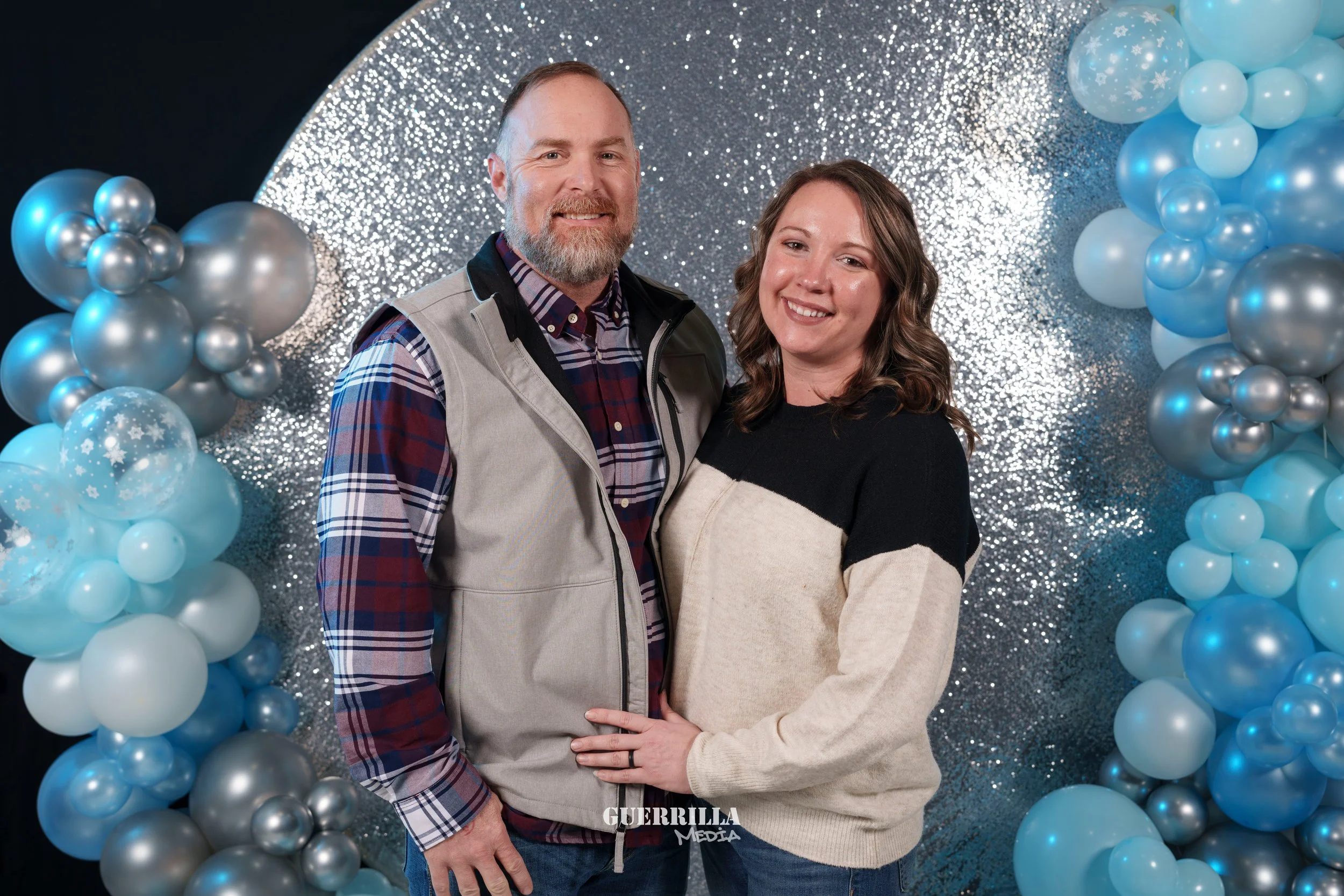 A smiling couple posing in front of a silver and blue balloon arch with snowflake decorations. The man has a beard and is wearing a plaid shirt with a gray vest, while the woman has wavy brown hair and is wearing a black and beige color-block sweater