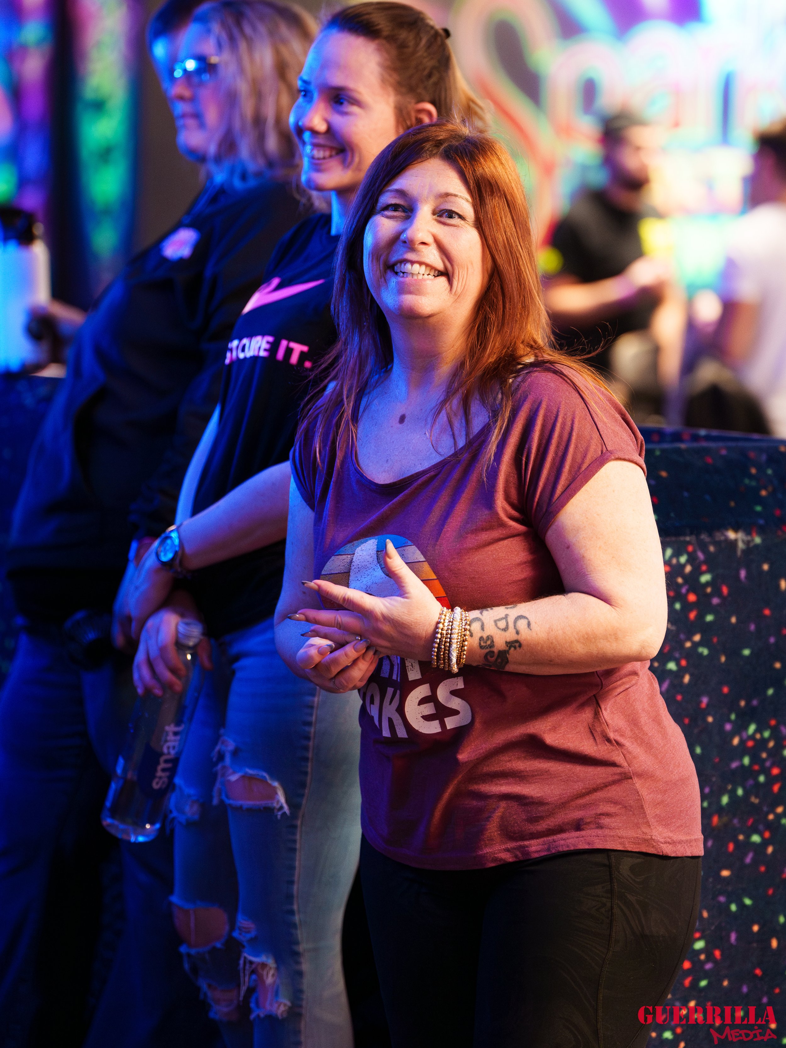 A group of women at a vibrant, neon-lit indoor venue, standing together and smiling. The woman in the foreground has red hair, tattoos on her arm, and is wearing a maroon t-shirt with a rainbow graphic and black pants. The background has a colorful, 