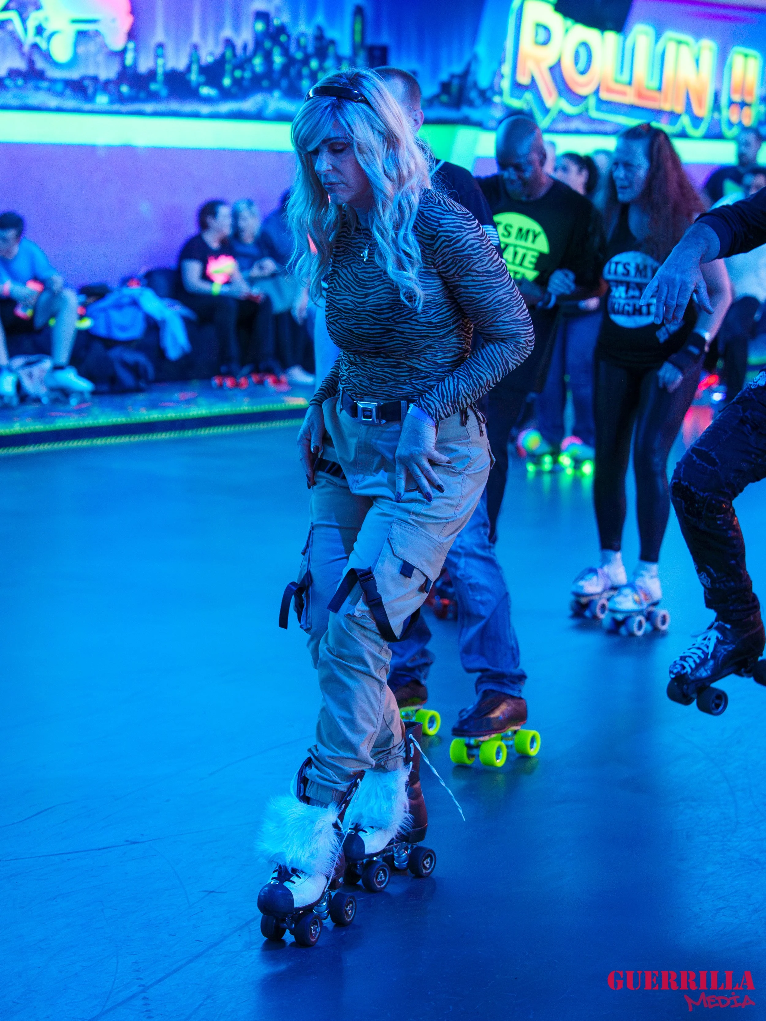 A woman roller skating at an indoor roller rink illuminated with neon and blacklight, with other skaters and neon-lit cityscape artwork in the background.