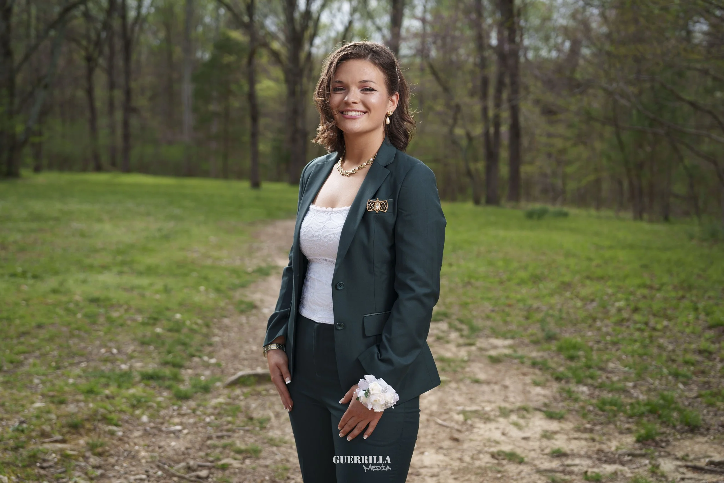 A woman in a dark green suit blazer and pants, white lace top, with gold jewelry and a white corsage, standing on a dirt path in a wooded area with green grass and trees, smiling at the camera.