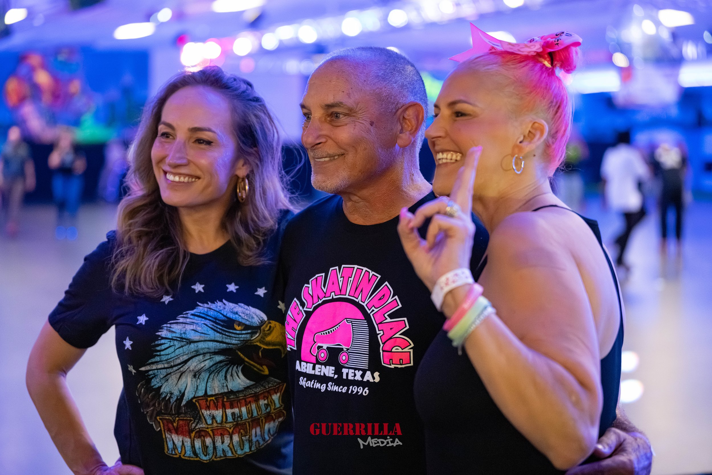 Three people smiling and posing together at an indoor roller skating rink, with colorful lights and people skating in the background.