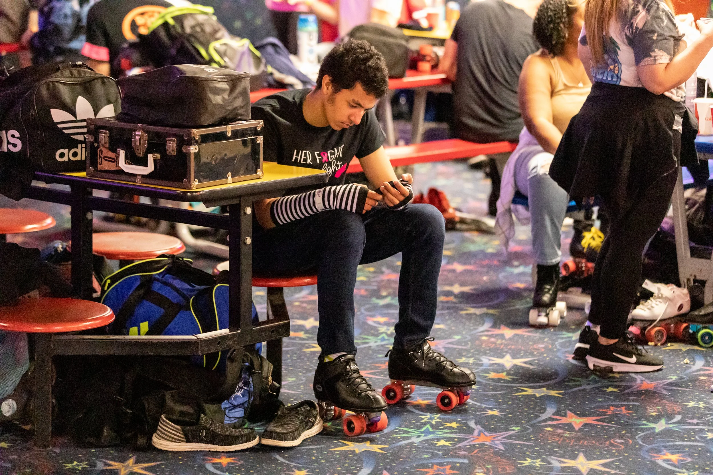 A young person sitting on a red bench in a roller skating rink, looking at their phone. They are wearing black roller skates, black pants, and a black t-shirt. There are backpacks and bags on the table and on the floor nearby, with other people in th