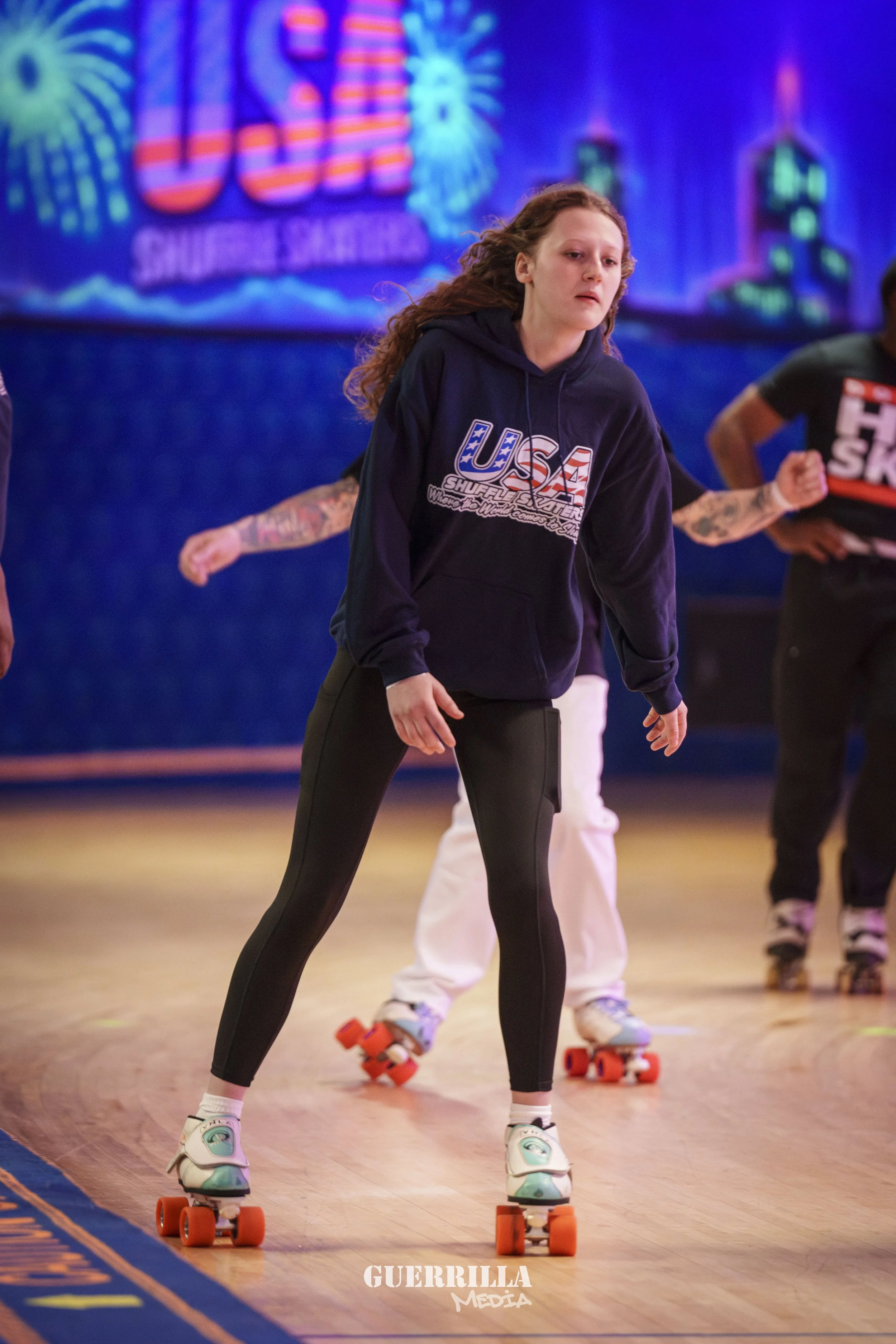 Young woman roller skating indoors with a focus on her wearing a navy hoodie with 'USA' on it, black leggings, and teal roller skates, participating in a skate event at a venue decorated with colorful neon lights and signs.