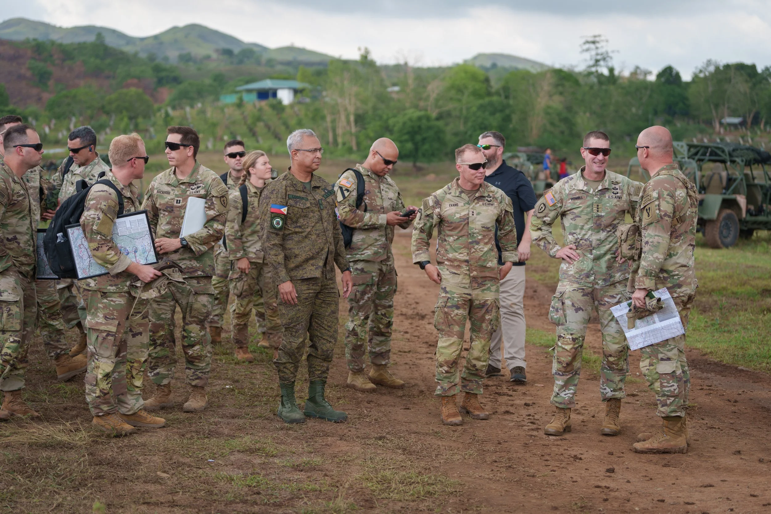 Group of military personnel in camouflage uniforms standing and talking outdoors on a dirt path, with green hills and trees in the background.