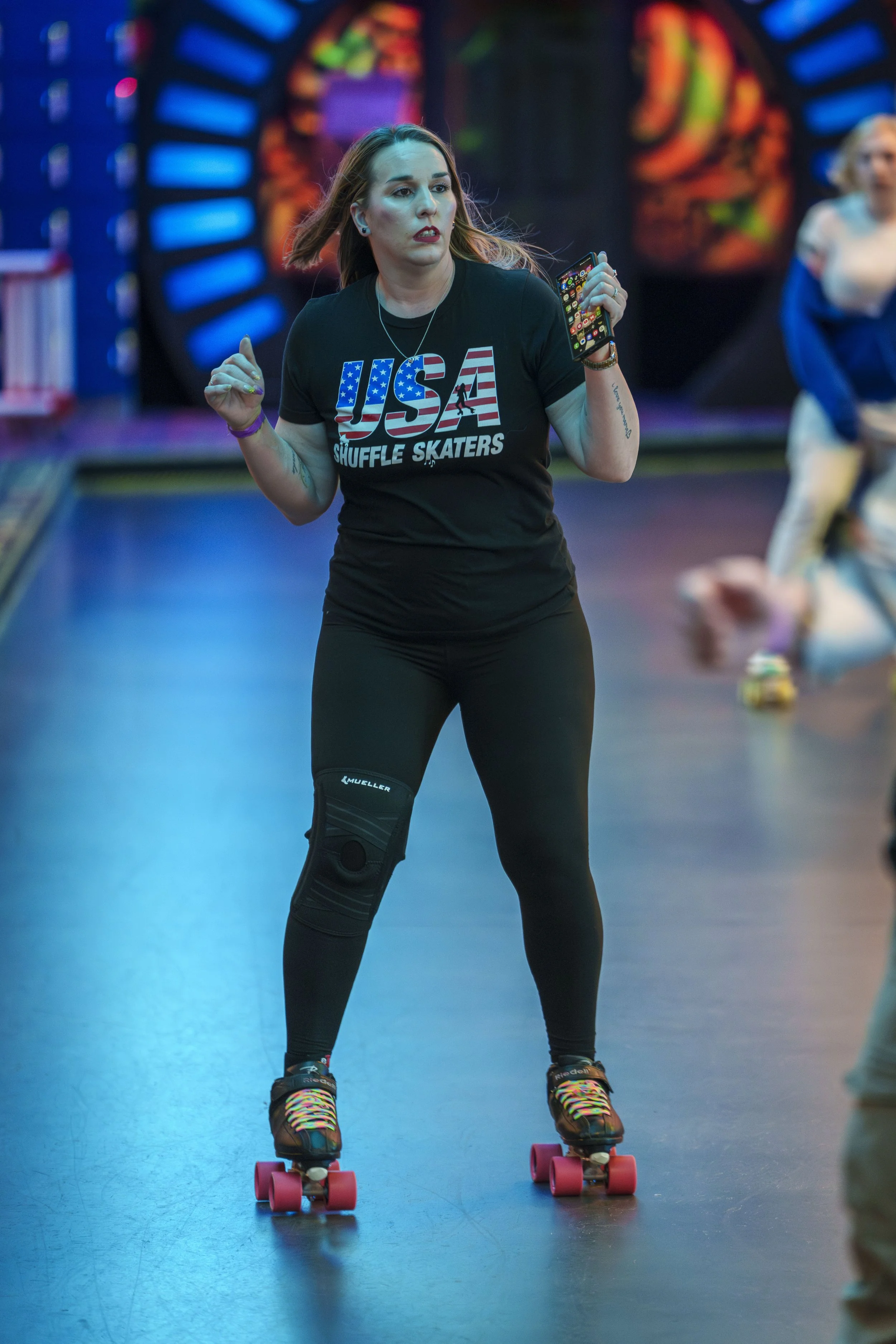 A woman dressed in a black T-shirt with 'USA Shuffle Skaters' printed on it, roller skating in an indoor arena with colorful lights and a blurred background, holding a smartphone in her right hand.